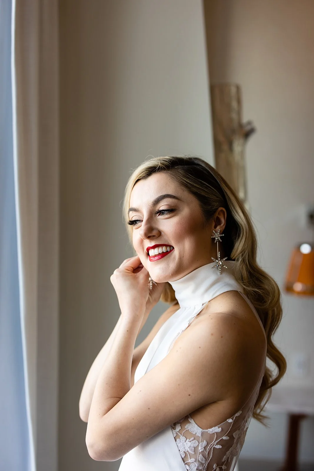 A young woman with blonde hair styled in loose waves, wearing a white dress with lace details on the shoulders, smiling and touching her face. She is wearing large star-shaped earrings with crystals, red lipstick, and has a joyful expression as she l
