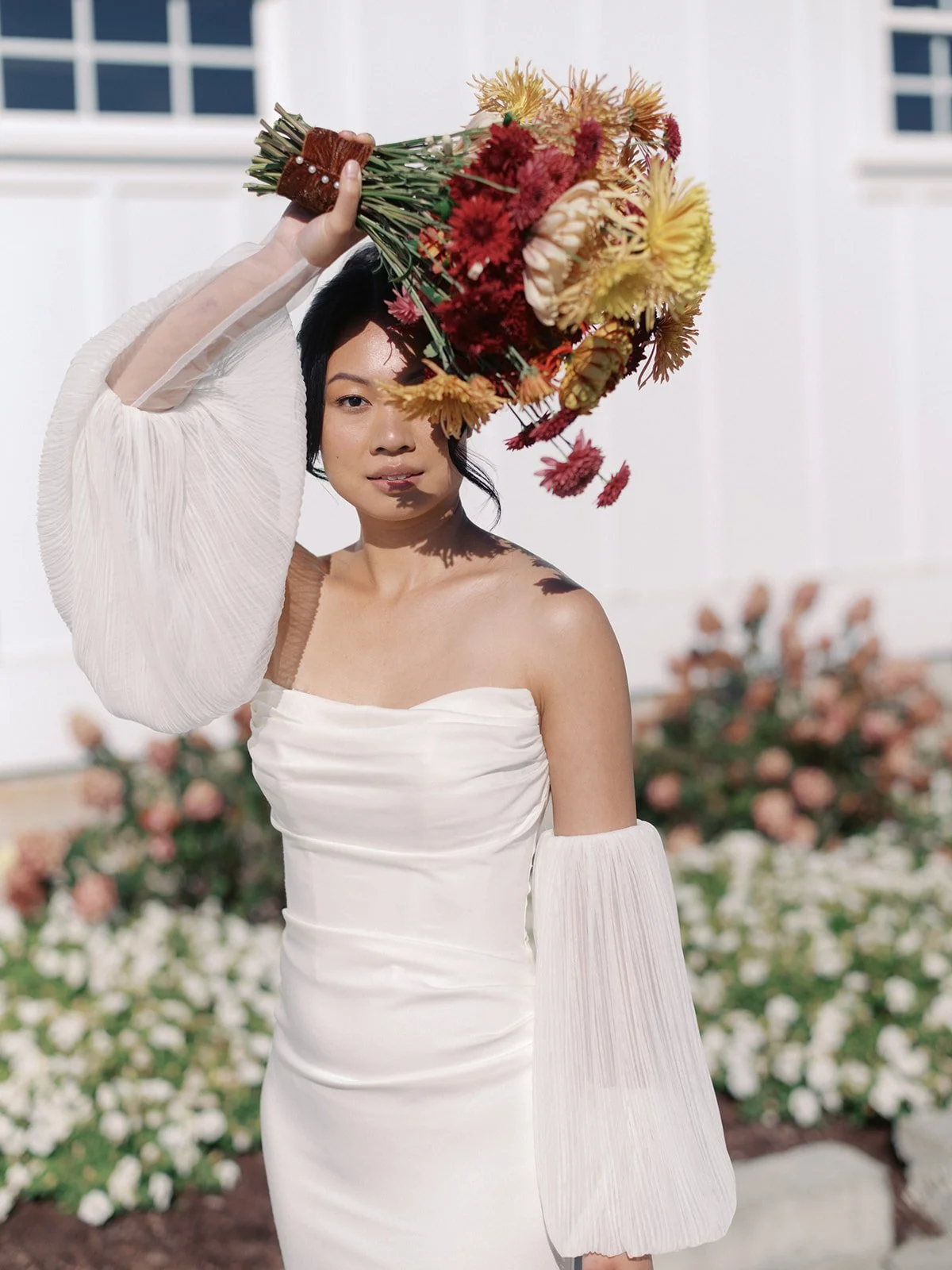 A woman wearing a white off-shoulder dress and sheer sleeves stands outdoors holding a bouquet of flowers above her head. The background shows a white wall and flower bed with pink and white flowers.