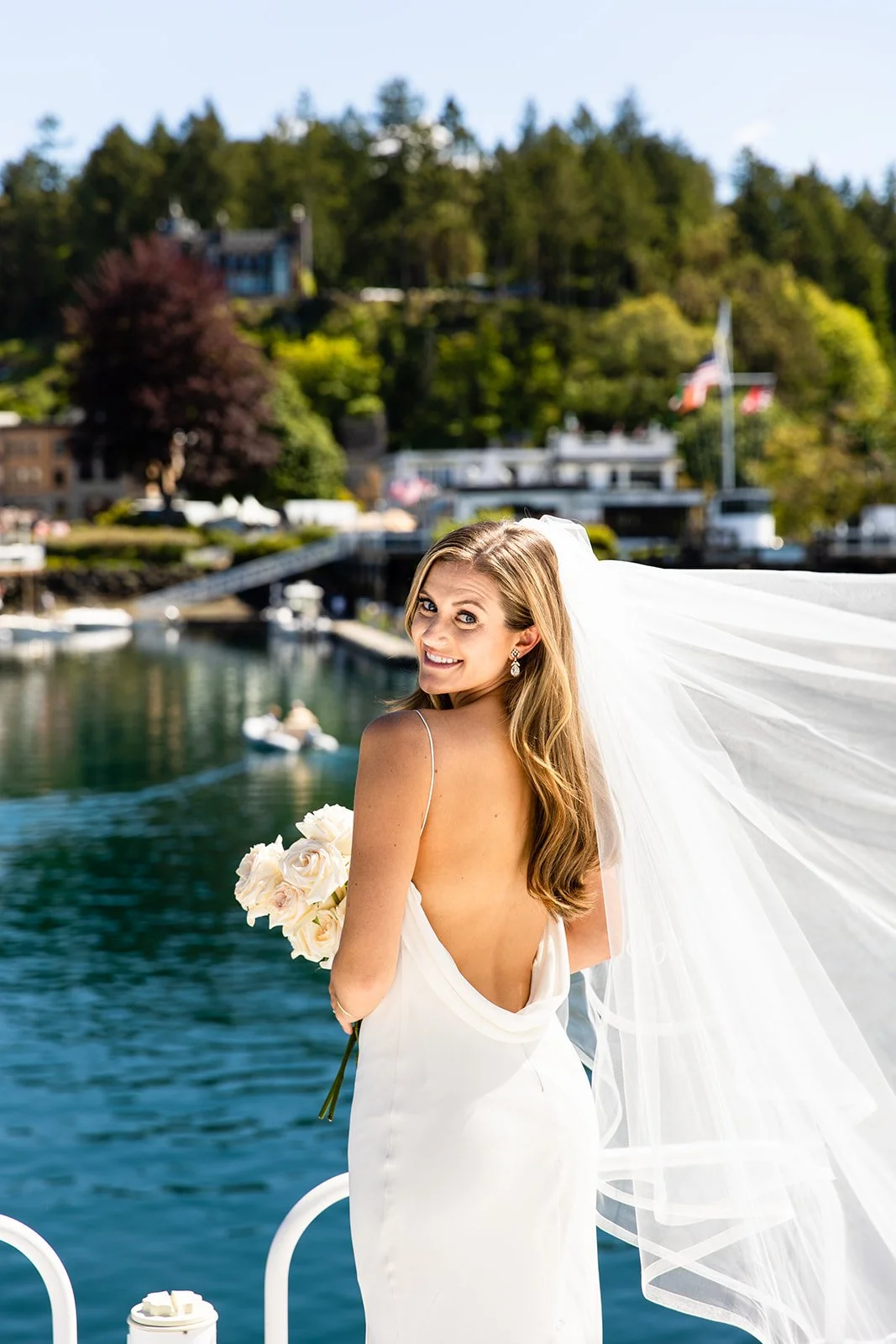 A bride in a white wedding dress holding a bouquet of white roses, standing outdoors by a body of water, smiling at the camera.