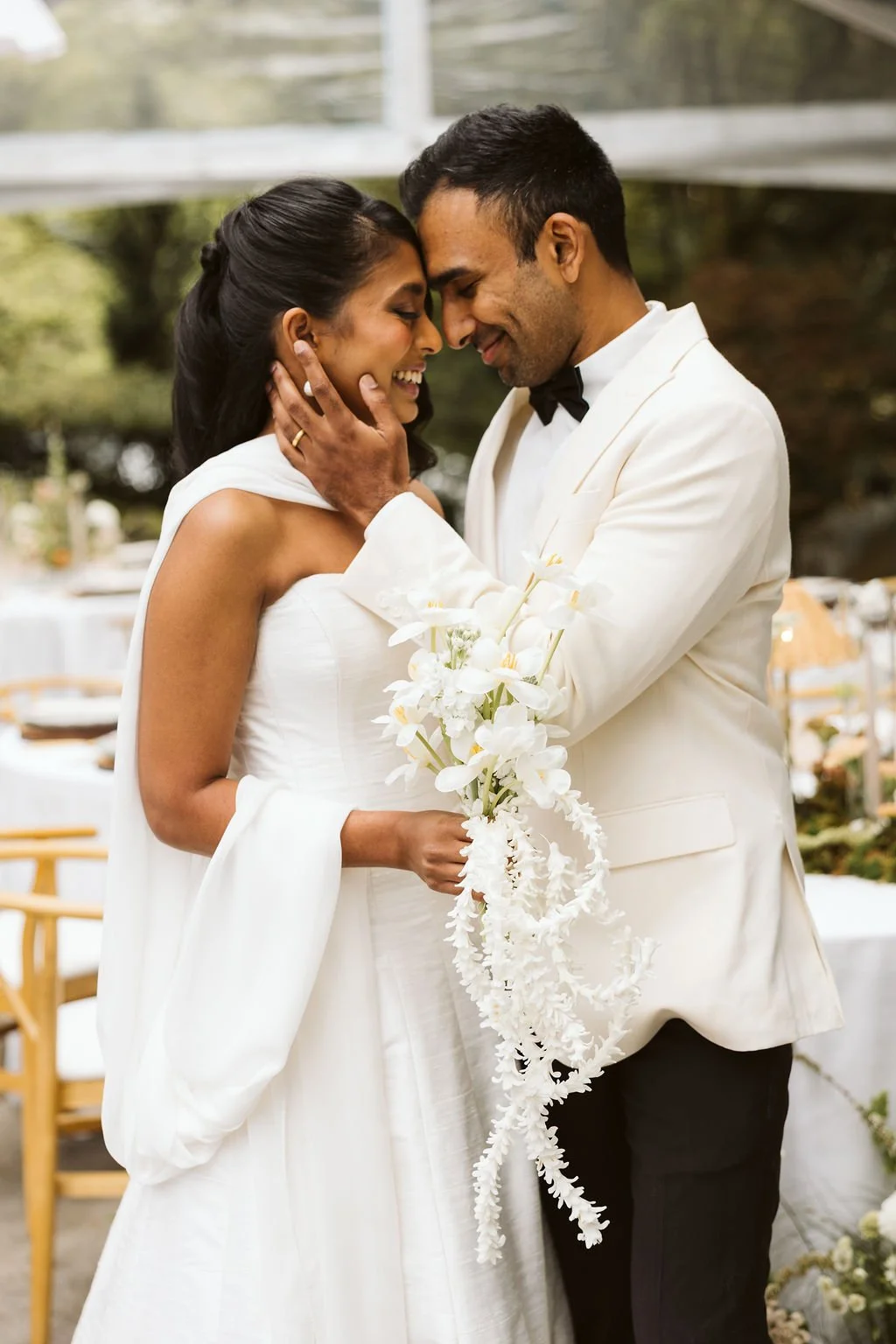 A couple at their wedding, dressed in white and black, sharing a close moment with foreheads touching and eyes closed, holding a bouquet of white flowers.