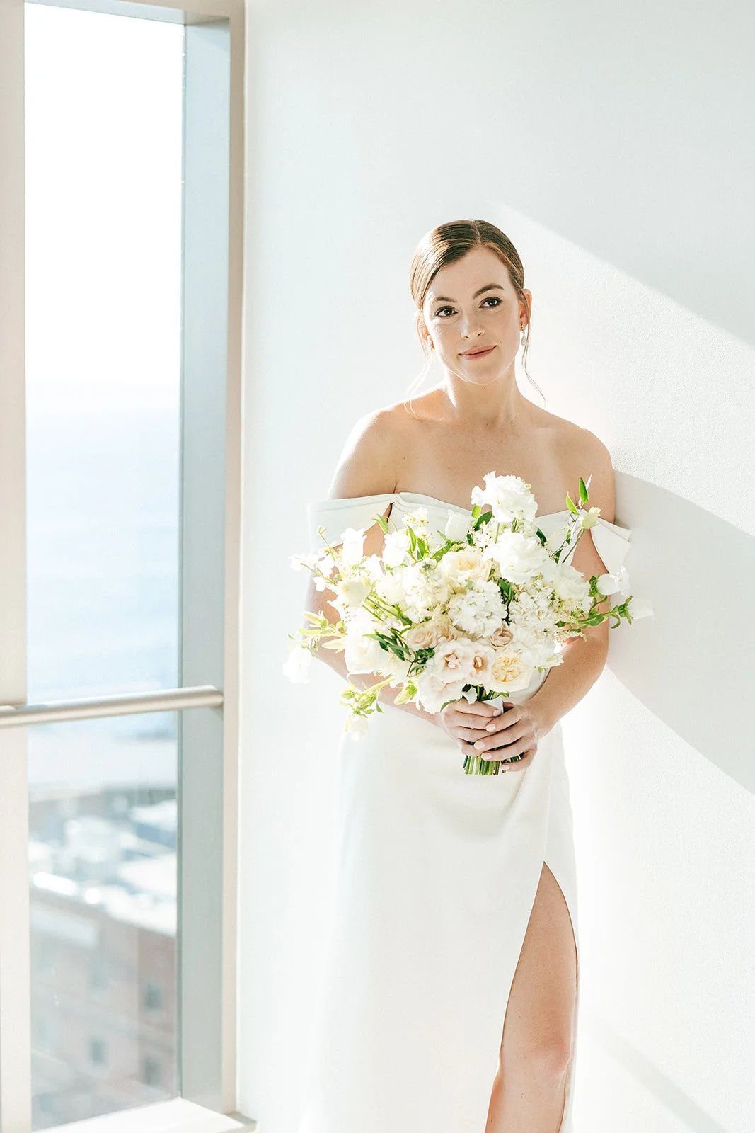 A woman in a white dress holding a large bouquet of white and light pink flowers, standing near a window with sunlight.