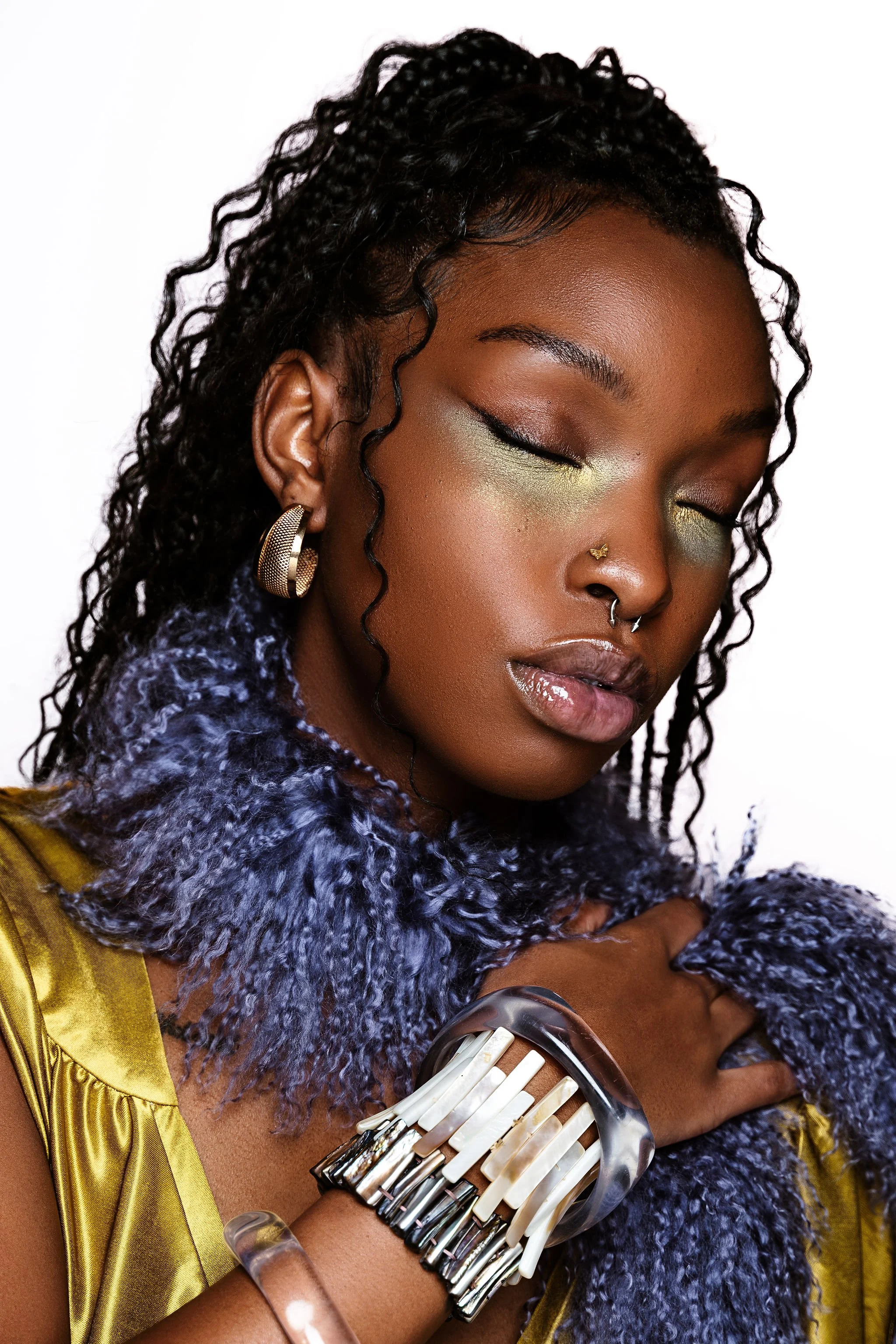 Close-up portrait of a woman with dark skin and curly black hair, wearing gold earrings, a metallic gold garment, and a dramatic makeup look with green eyeshadow, gold highlighter, and a septum piercing. She is holding a large, artistic bracelet.