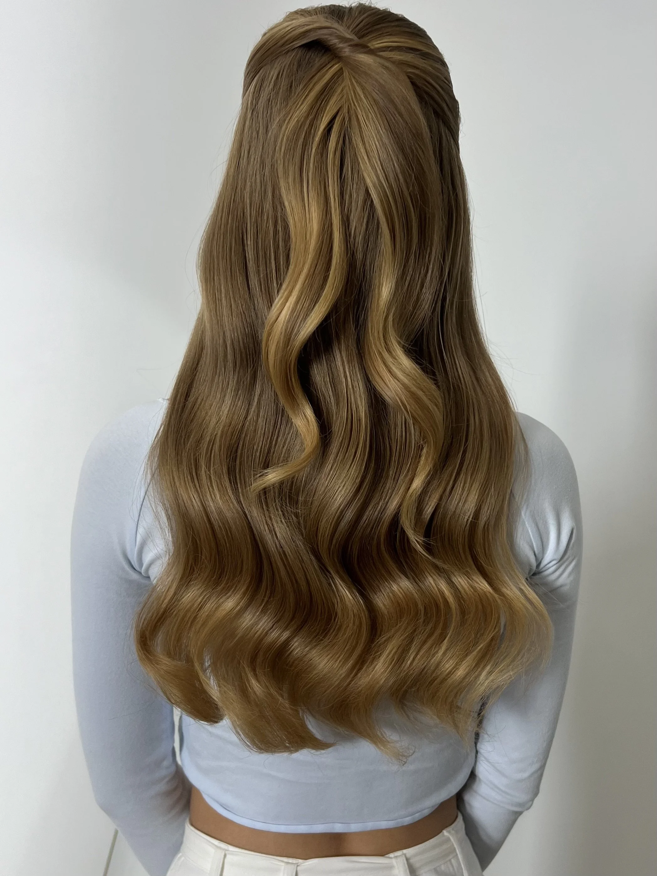 A woman with long, wavy, blonde hair seen from behind, wearing a light gray long sleeve top and white pants.