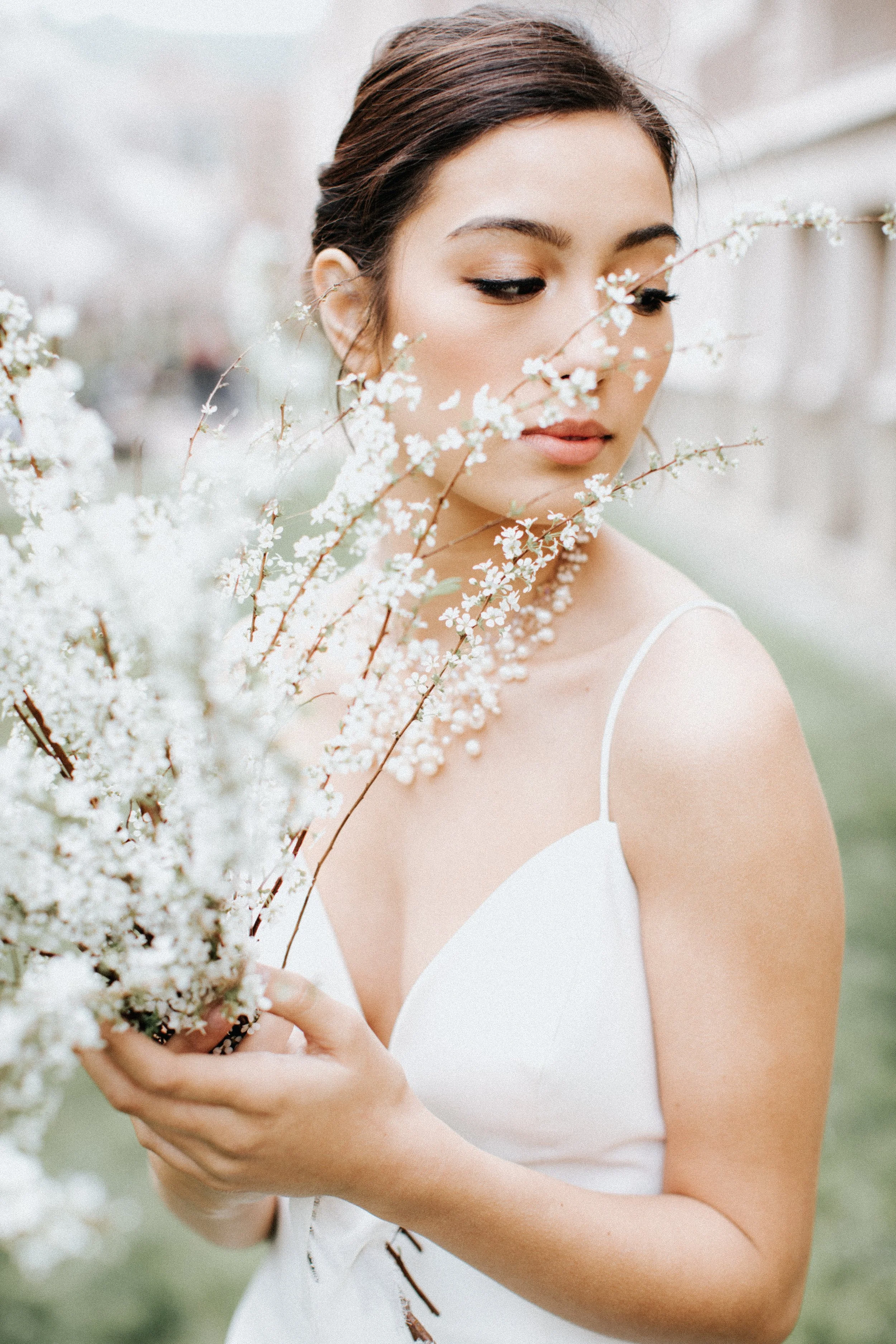 A young woman with brown hair in an elegant updo, wearing a white dress and necklace, holding a bouquet of white flowers with small blossoms, outdoors with a blurred background.