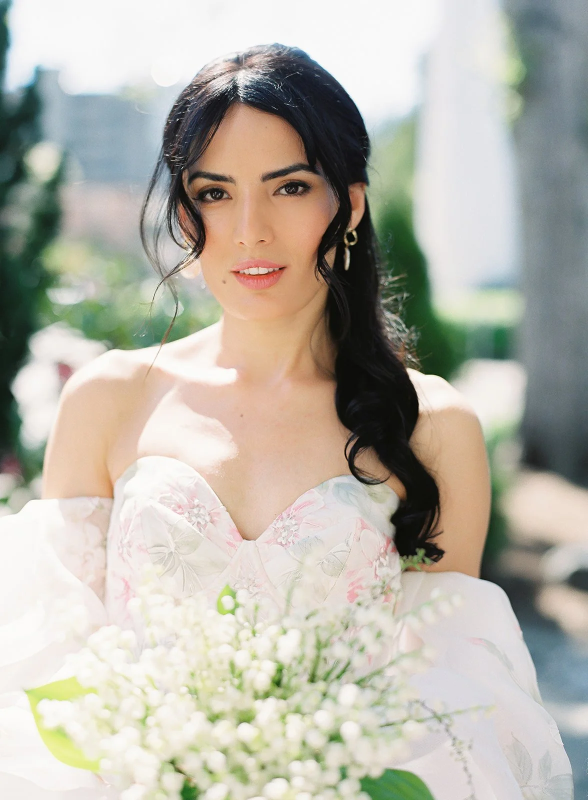 A woman with long dark hair in soft waves, wearing earrings and a floral off-the-shoulder dress, is outdoors with greenery in the background, holding a bouquet of white flowers.