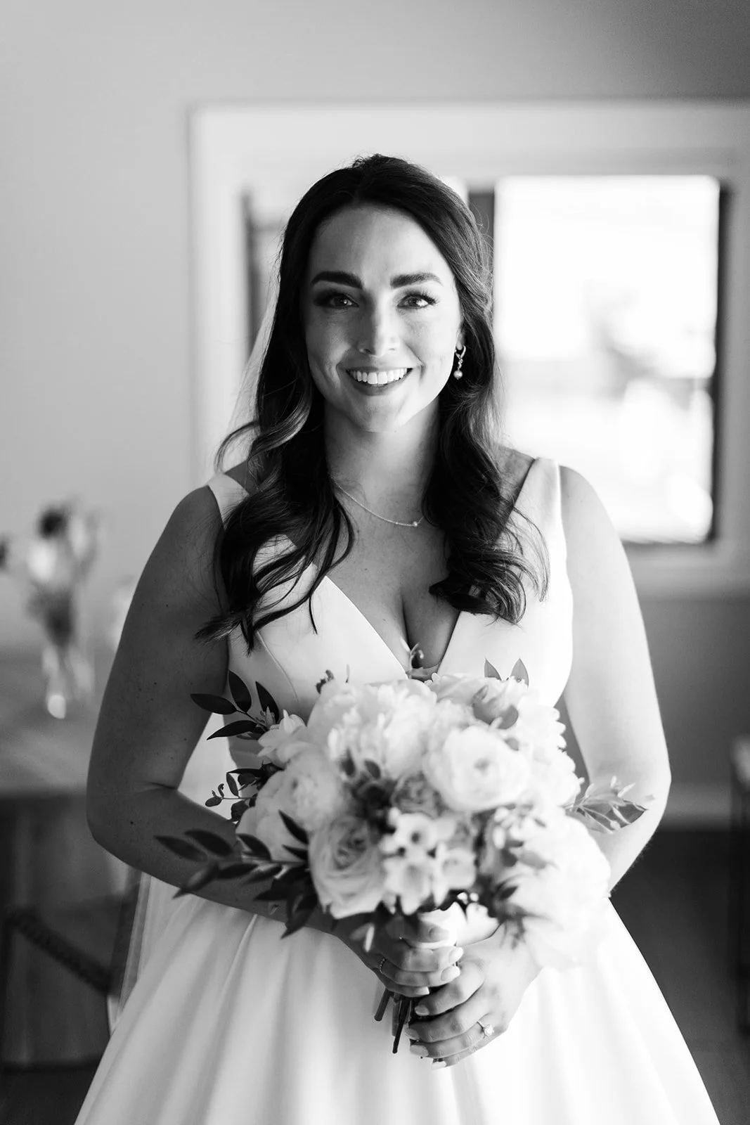 Black and white photo of a smiling woman in a wedding dress holding a bouquet of flowers, standing indoors near a window.