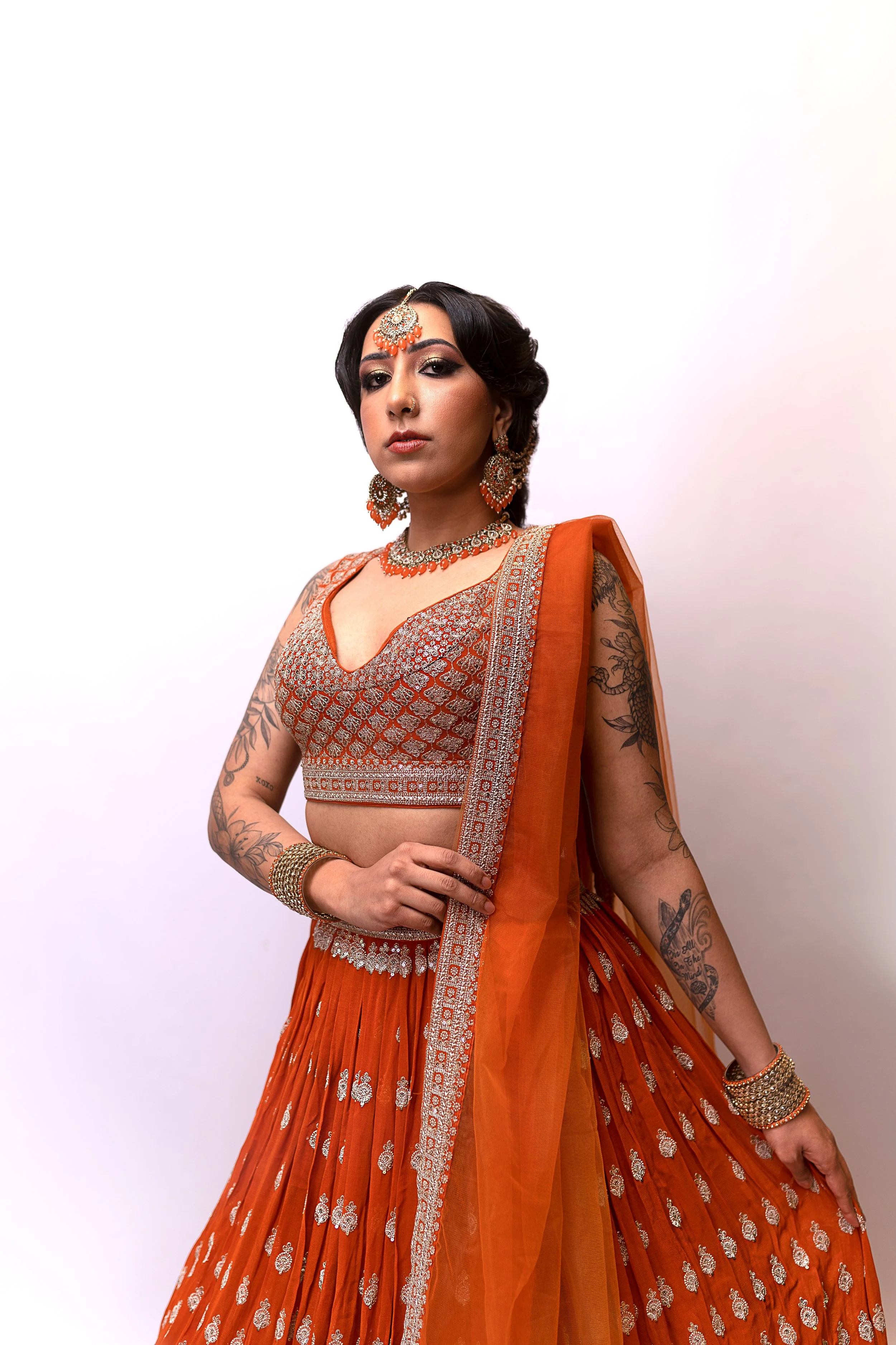 A woman with dark hair and tattoos wearing traditional orange and gold Indian attire and jewelry, standing against a white background.