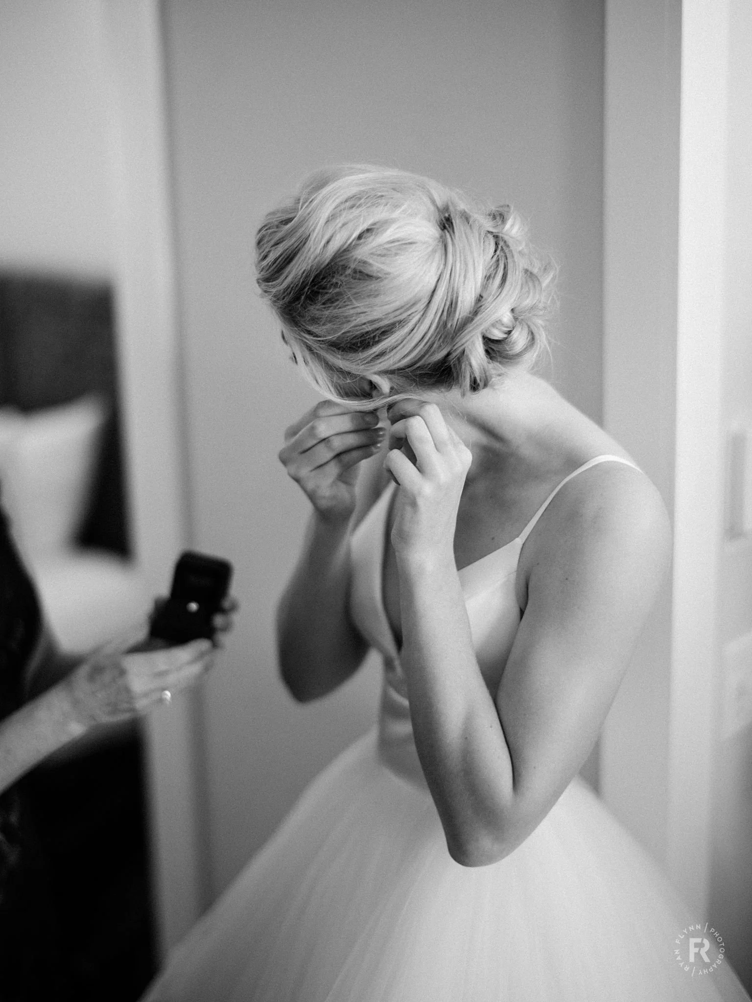 Black and white photo of a woman with curly blonde hair adjusting her earring in front of a mirror, wearing a sleeveless top.