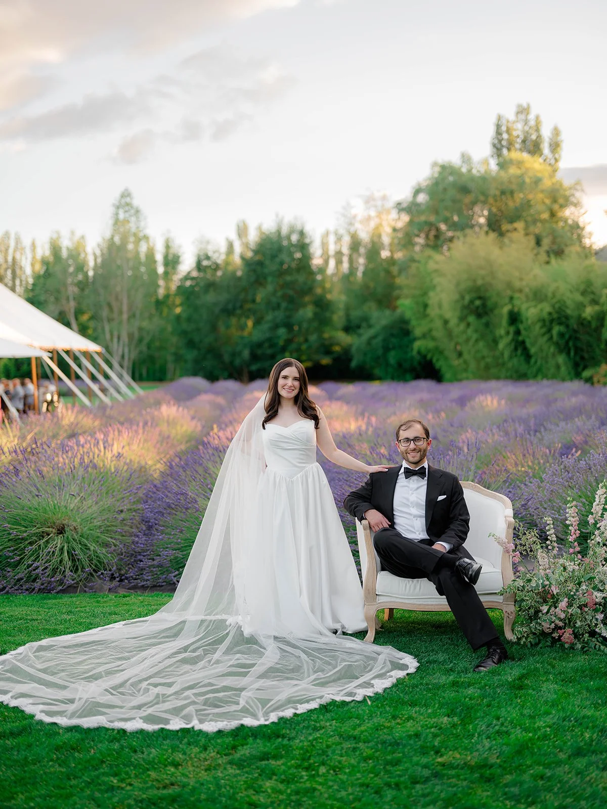 A bride and groom posing outdoors in a garden with purple flowers. The bride is in a white wedding dress, with a veil, standing and resting her hand on the groom's shoulder. The groom is seated on a white sofa, wearing a black tuxedo, glasses, and sm