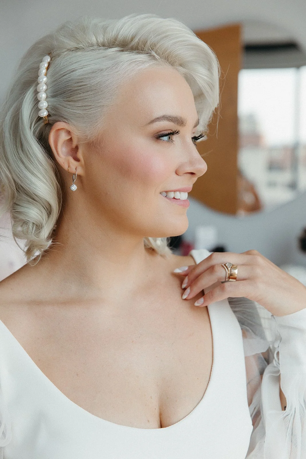 A smiling woman with platinum blonde hair styled in loose waves, wearing pearl hair accessories, diamond earrings, and a white top, indoors with a window in the background.