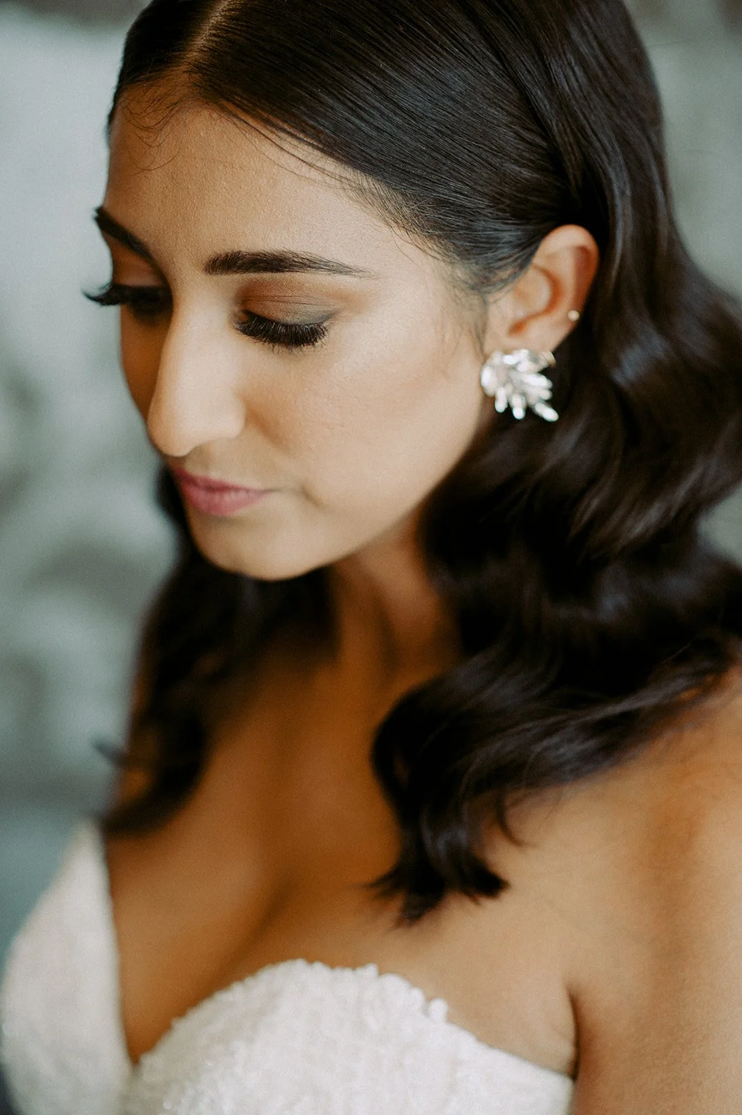 Close-up of a woman with dark brown hair styled in loose waves, wearing a strapless white lace dress and silver earrings, looking downward with her eyes closed.
