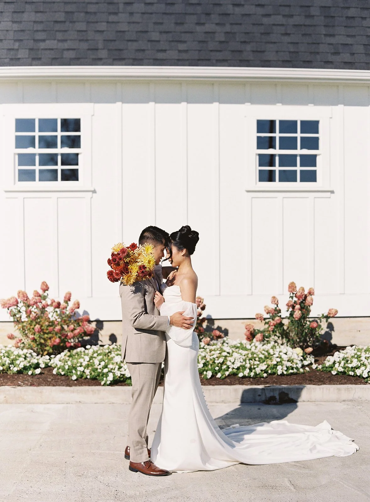 A bride and groom standing close together outdoors, touching foreheads, with the groom holding a bouquet of red and yellow flowers, in front of a white building with two windows and surrounded by pink and white flowers.