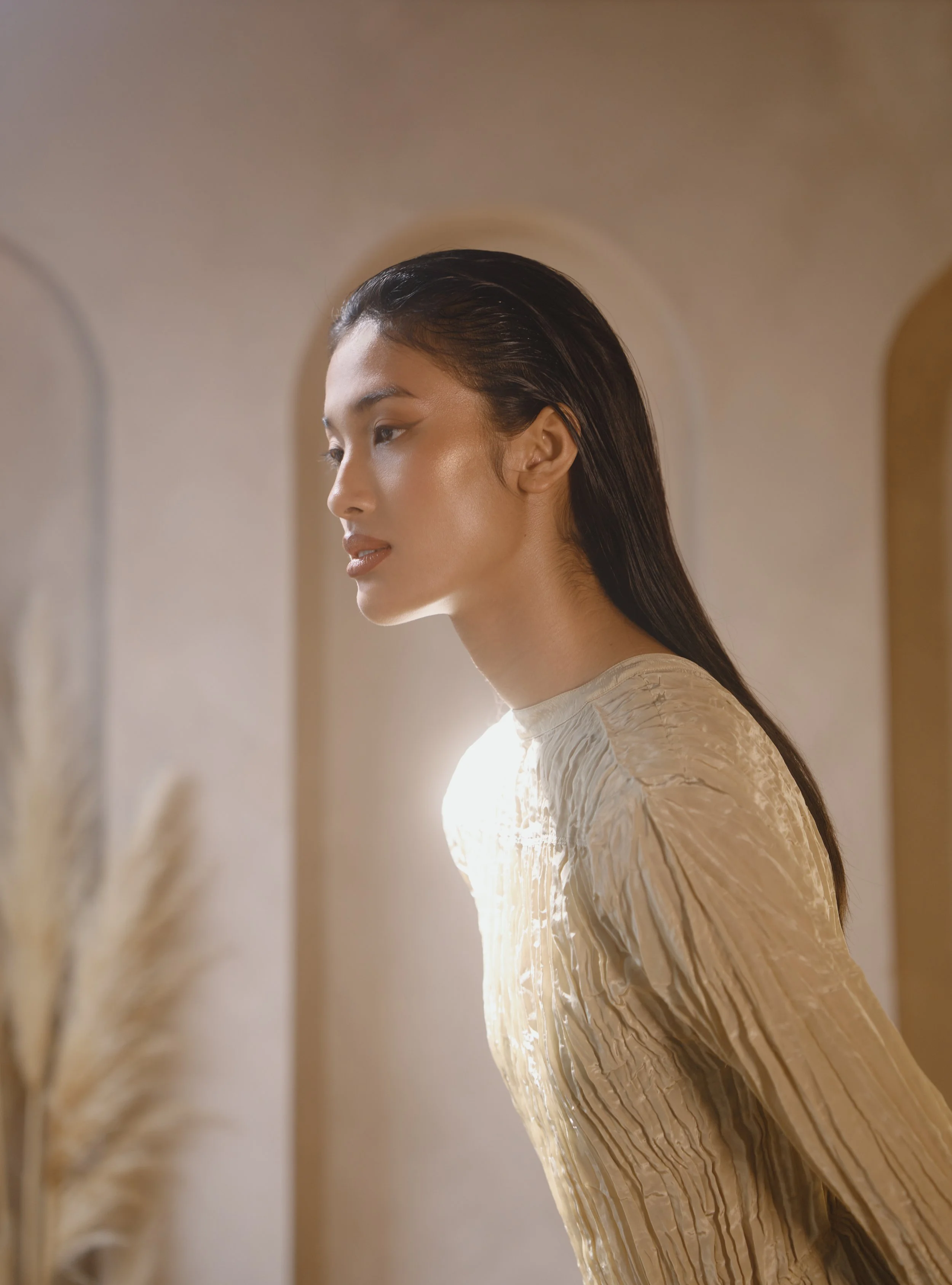 A woman with long dark hair and light skin is seen in profile, wearing a cream-colored, textured long-sleeve top. She is standing in front of a mirror with soft, warm lighting.