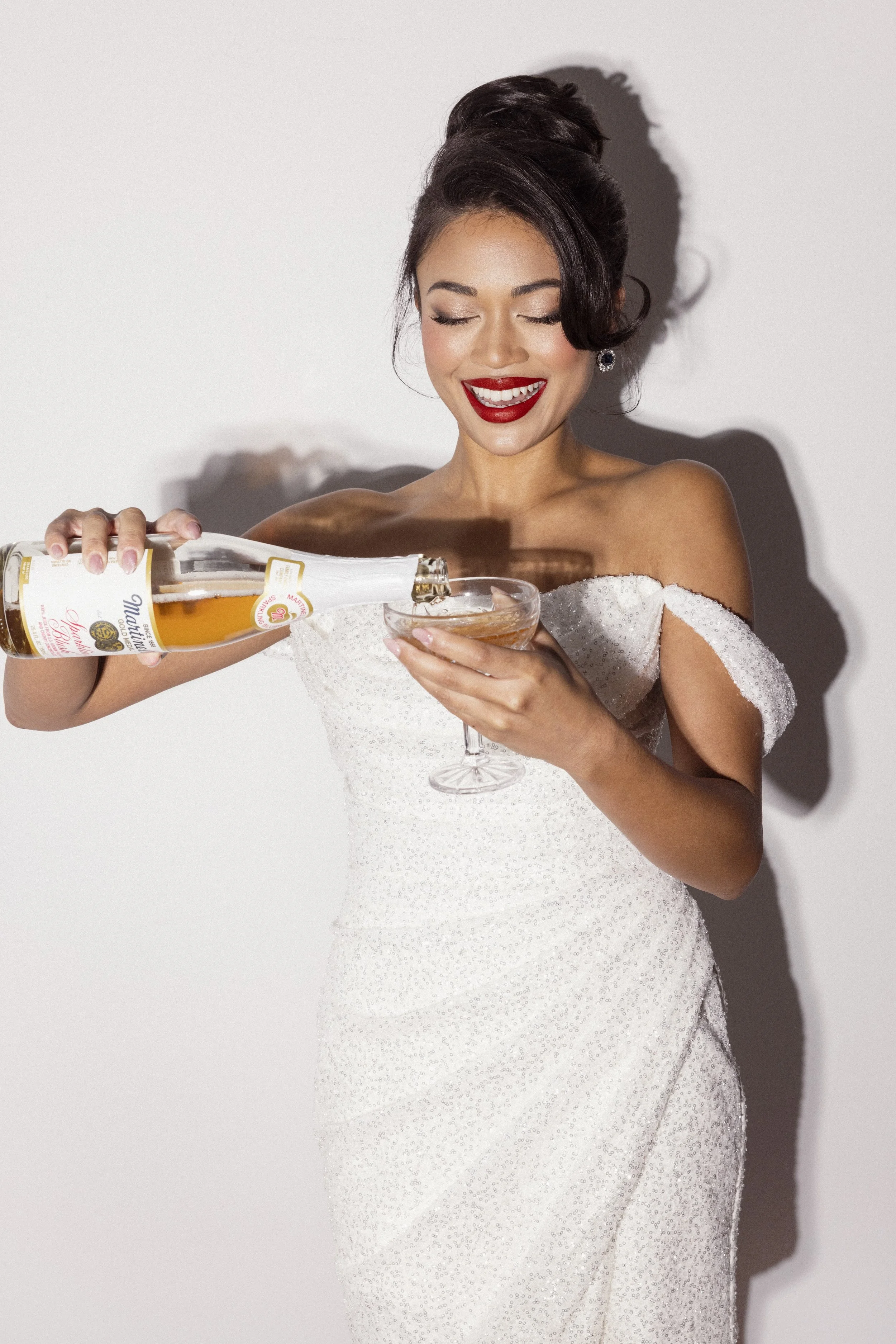 A woman in a white dress with off-shoulder sleeves is smiling and pouring champagne into a glass. She has dark hair styled in an elegant updo, red lipstick, and is wearing earrings. The background is plain and white, with a shadow cast on the wall.