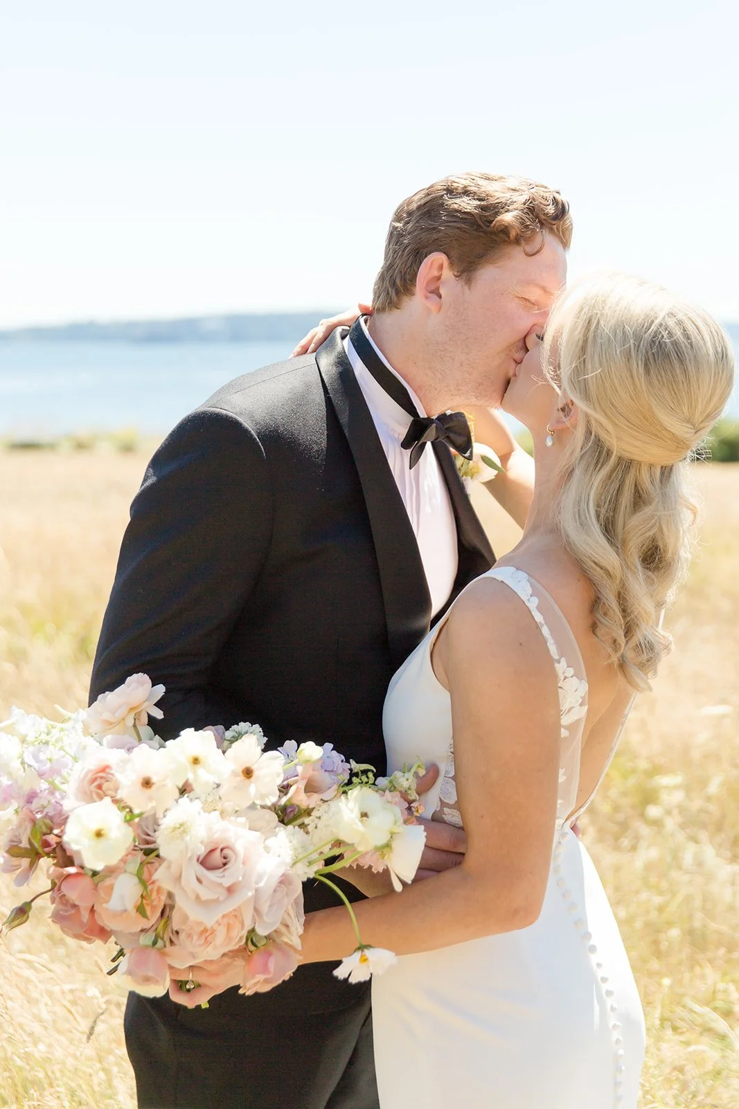 Bride and groom share a kiss outdoors on a sunny day, with a field and water in the background; the bride holds a bouquet of pink and white flowers.