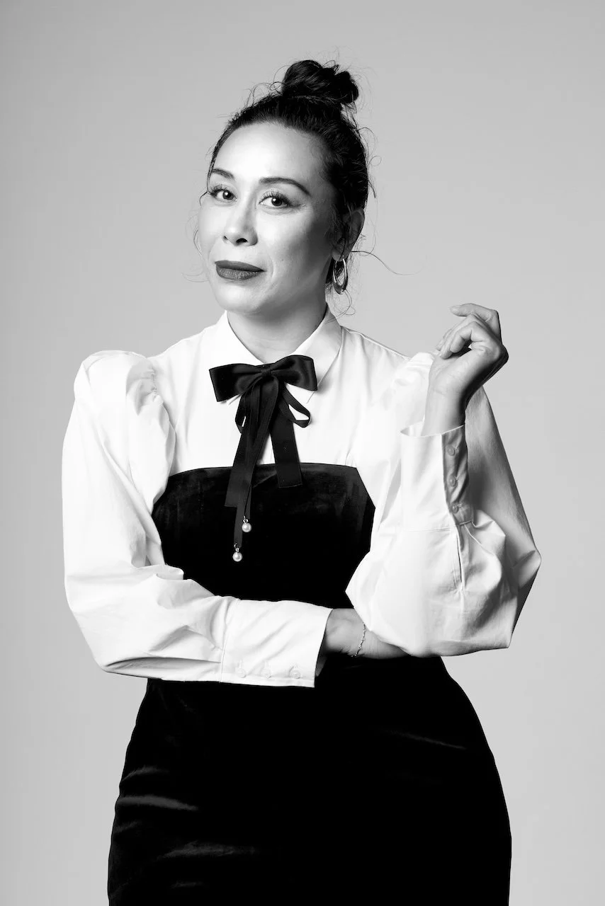 A woman with dark hair in a bun, wearing a white blouse with puffed sleeves, a black pinstripe vest, and a black bow tie, posing against a light gray background.