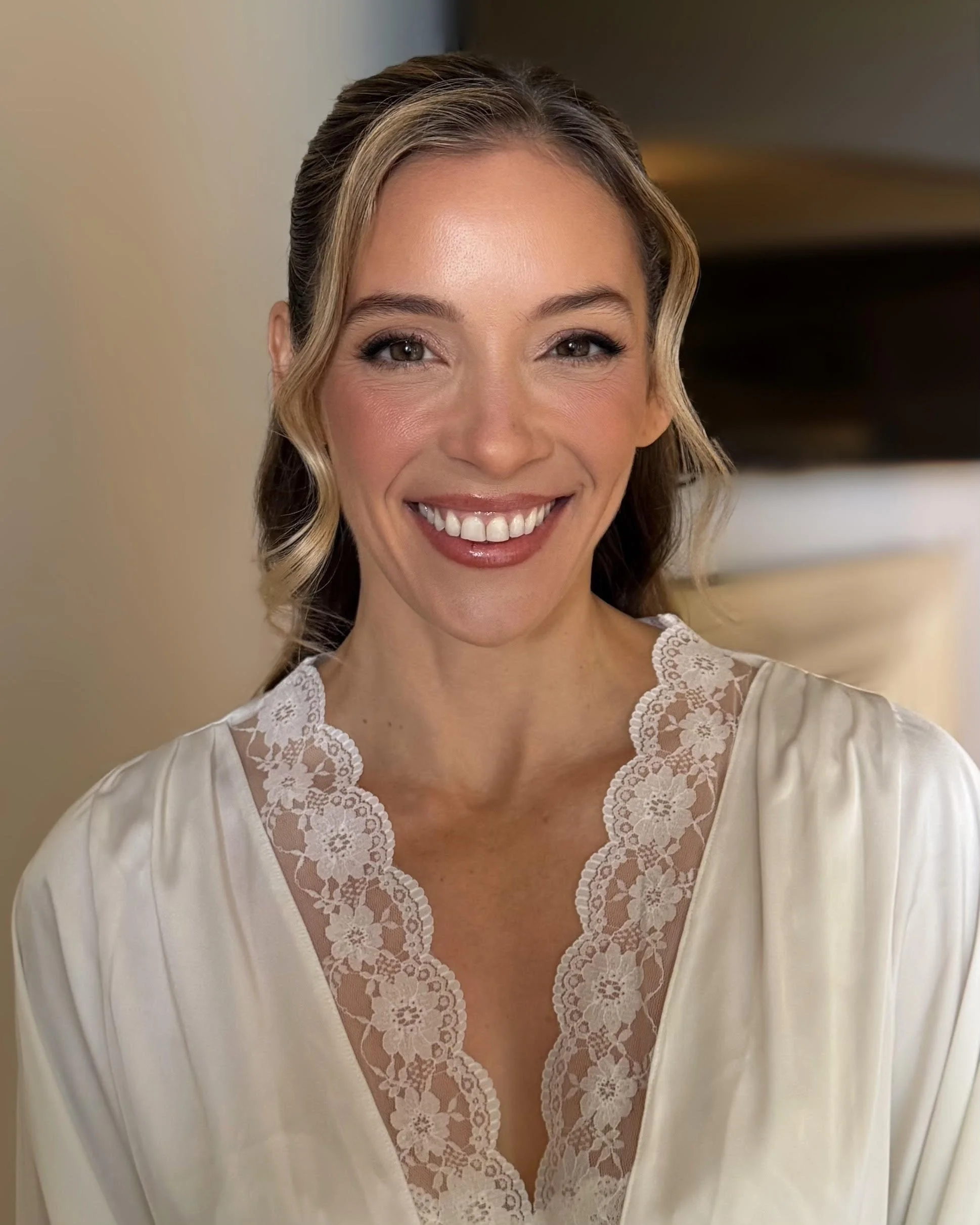 Close-up of a woman with wavy blonde hair and brown eyes, smiling, wearing a white lace-trimmed top.