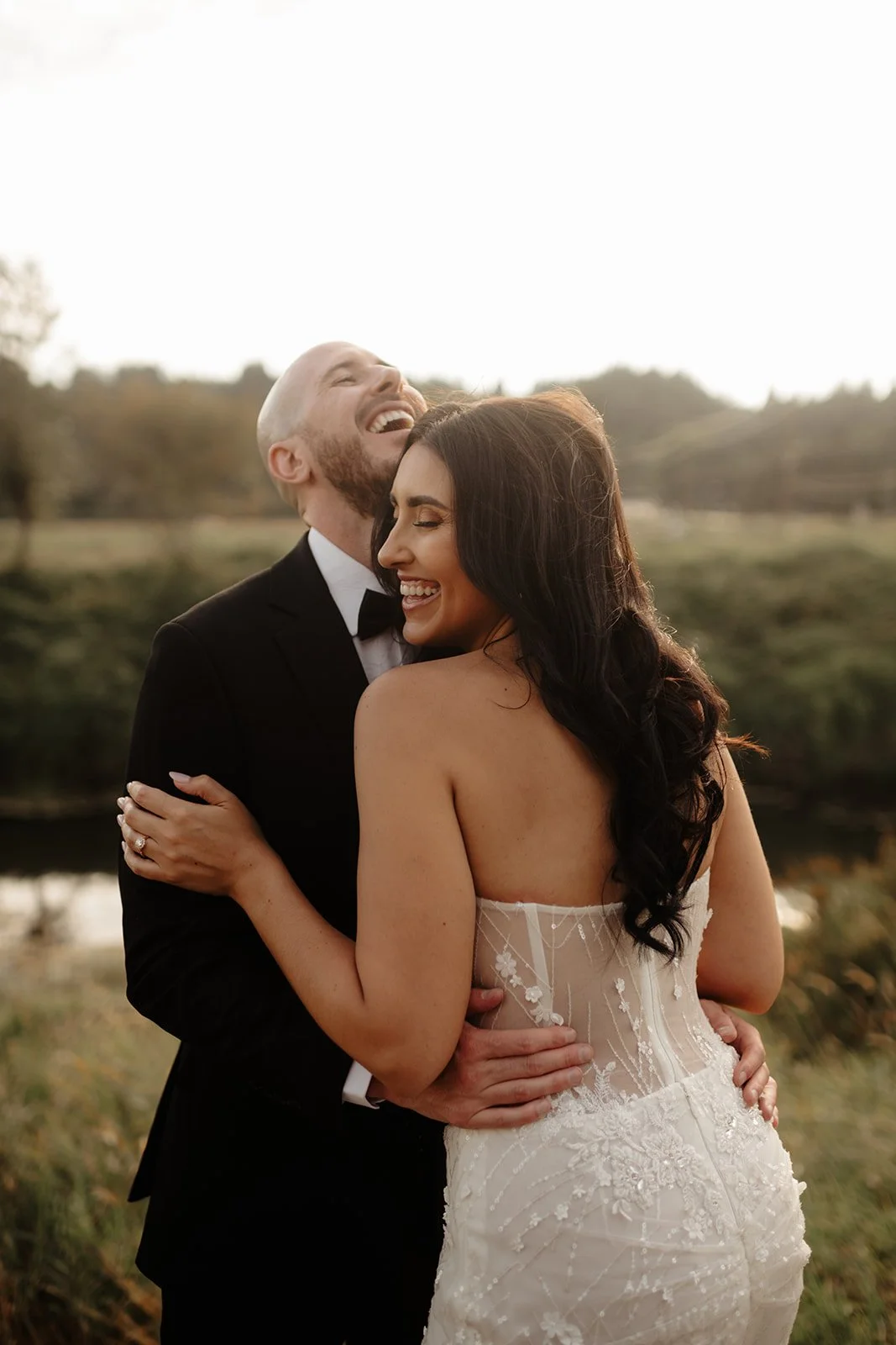 A newlywed couple dressed in wedding attire sharing a joyful embrace outdoors during sunset, with trees and a river in the background.