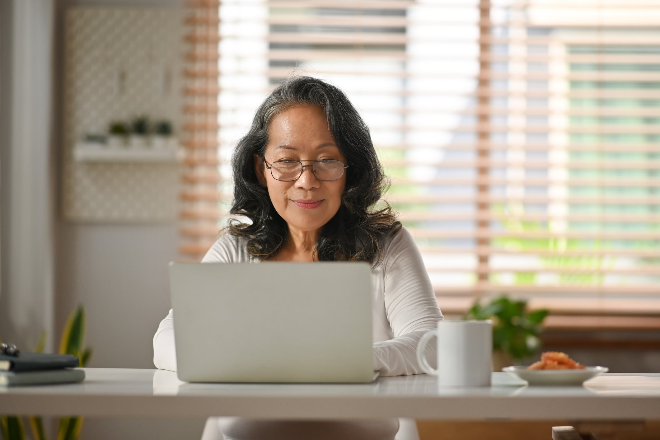 An elderly woman wearing glasses sitting at a table with a laptop in front of her, a white mug, and a plate with food, in a bright room with window blinds in the background.