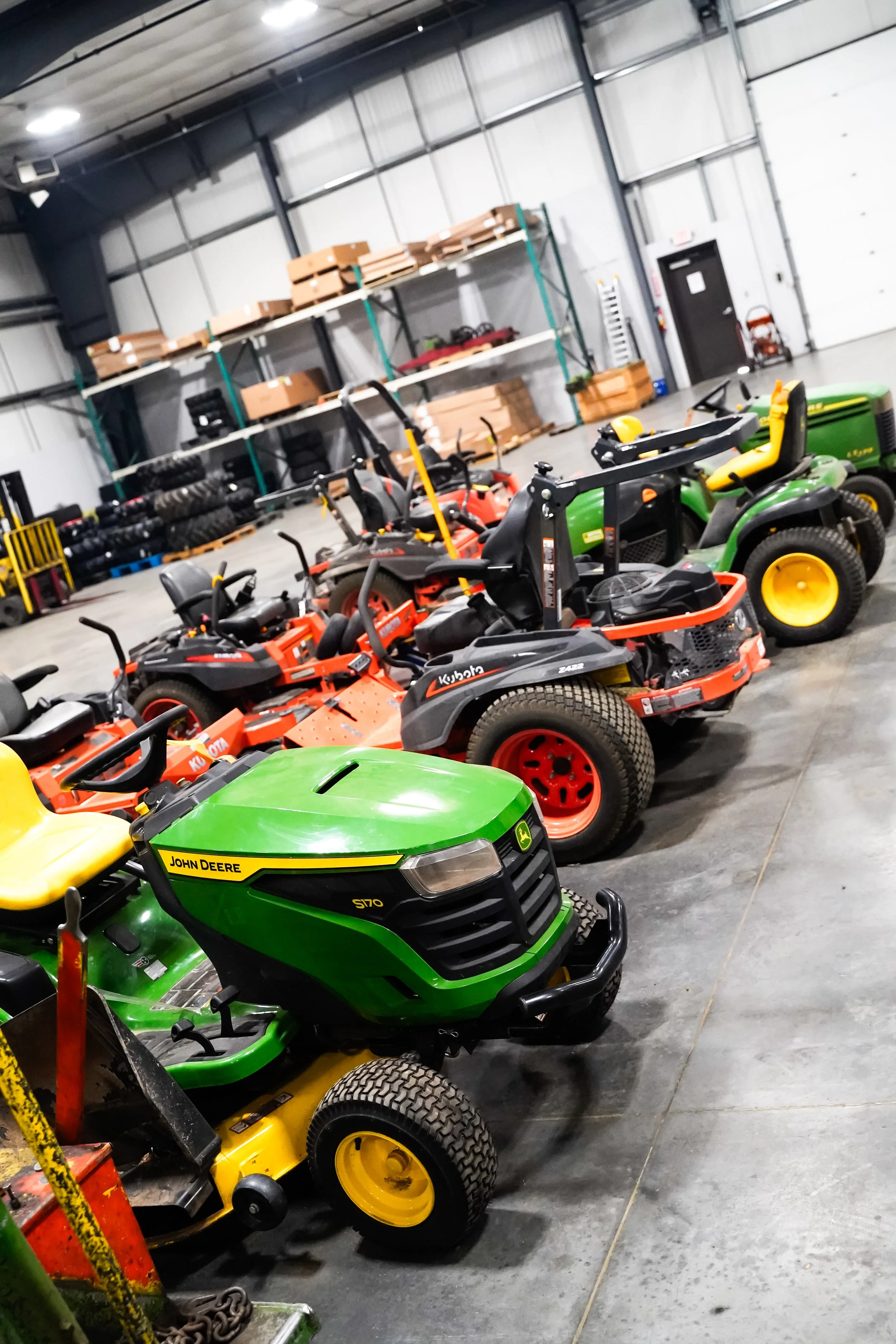 Indoor storage area with several riding lawn mowers, including a green John Deere, and shelving with boxes and equipment in the background.