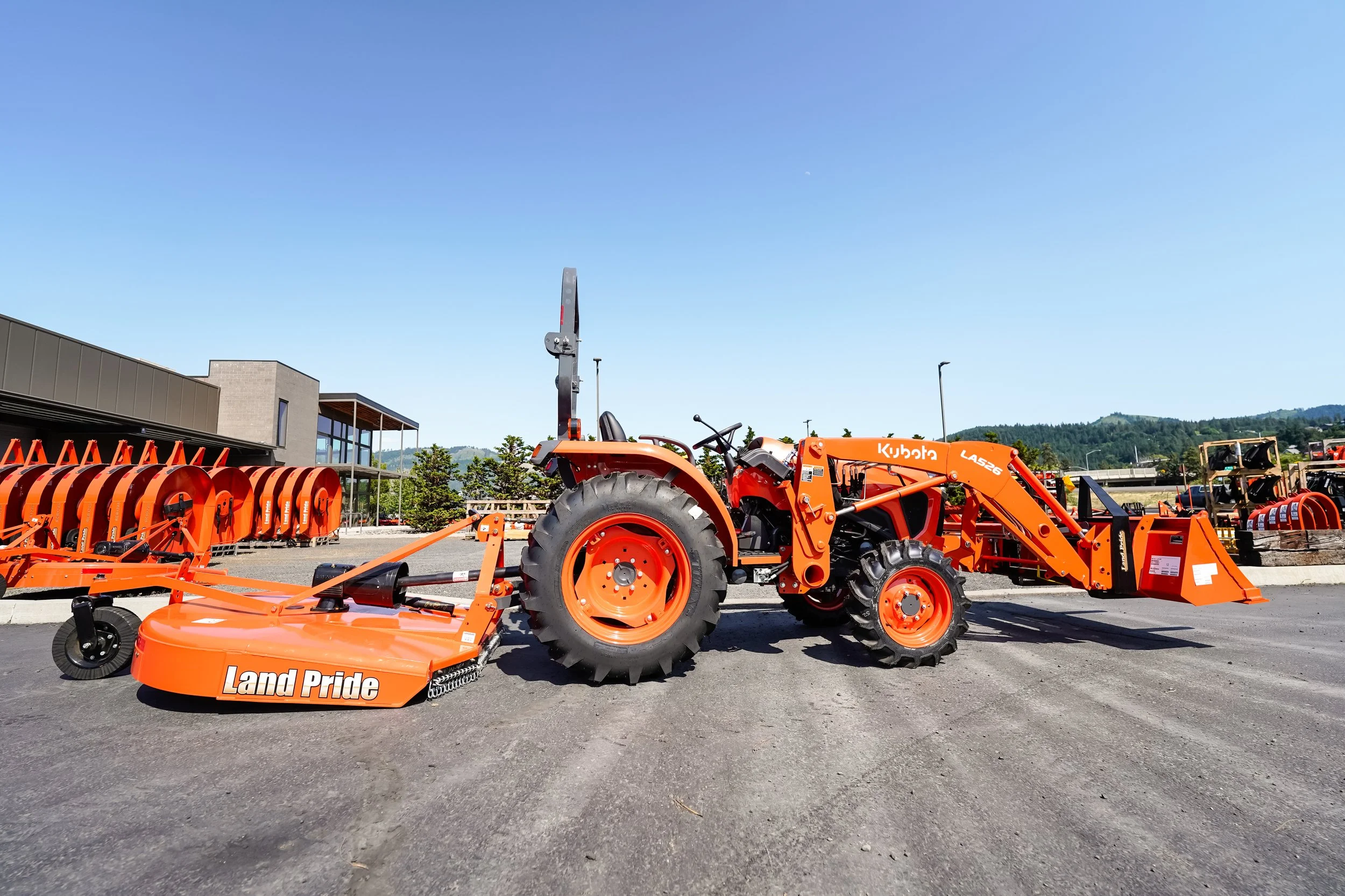 Orange Kubota tractor with attachments parked outdoors on asphalt, with a building, trees, and mountains in the background.