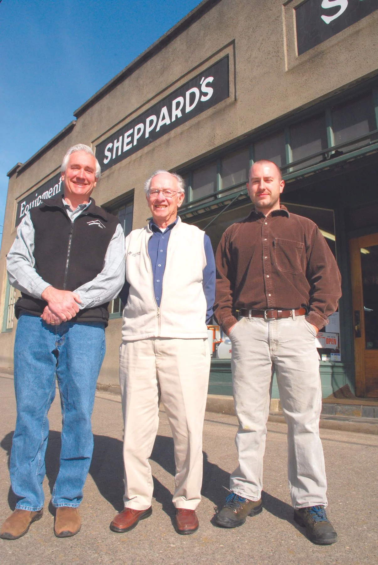 Three men standing outside a store called Sheppard's, smiling. The building has a tan exterior, and the store sign is black with white text. The man on the left wears a black vest over a checkered shirt, blue jeans, and tan boots. The man in the center wears glasses, a white vest, a blue shirt, cream-colored pants, and brown shoes. The man on the right wears a brown shirt, light-colored pants, and dark shoes. The sky is clear and blue.