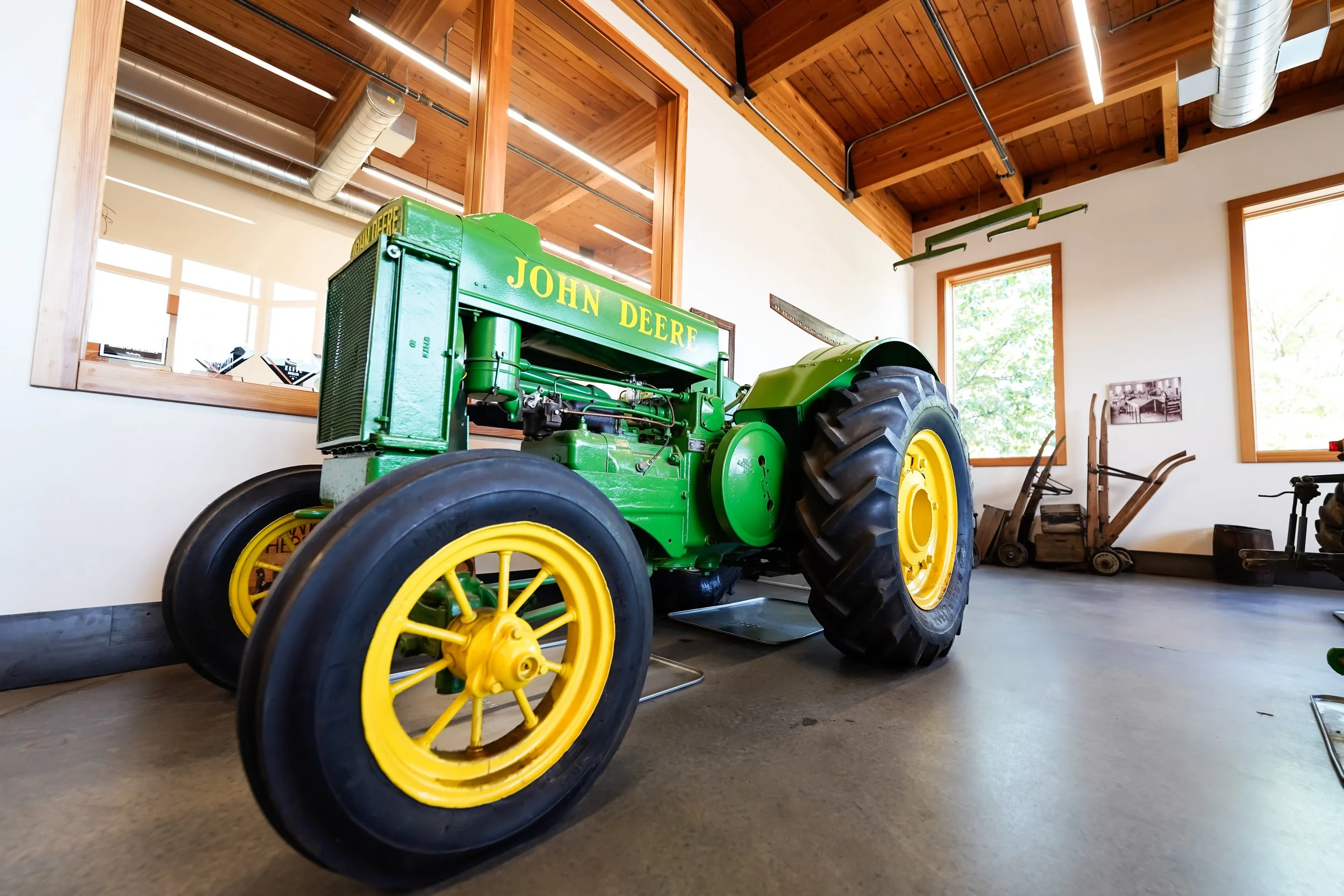 A vintage green John Deere tractor with yellow wheels inside a room with wooden ceiling beams and large windows, reflecting greenery outside.