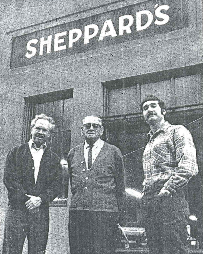 Black and white photo of three men standing in front of a building with a sign that reads 'Sheppard's.' The man on the left is smiling, wearing glasses, a dark sweater over a shirt. The man in the middle, with glasses and a beard, is wearing a blazer and tie. The man on the right has dark hair, is wearing a plaid shirt, and has his hands in his pockets.