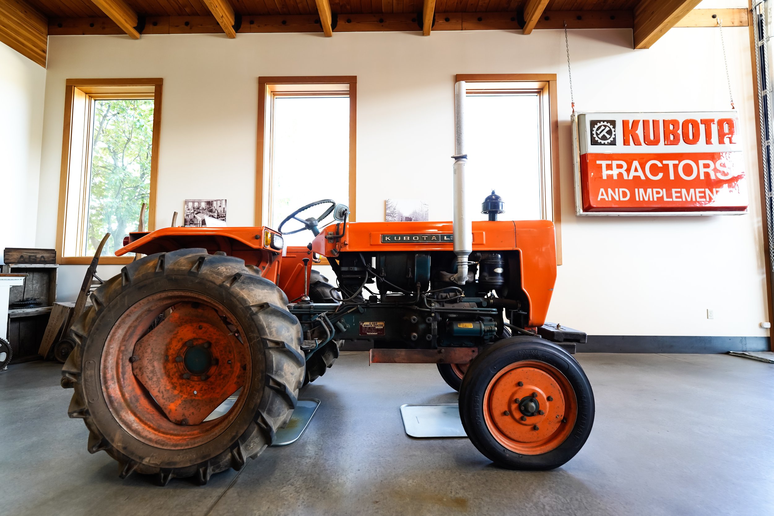 Orange Kubota tractor inside a room with three windows and a wooden ceiling. A sign on the wall reads 'KUBOTA TRACTORS AND IMPLEMENT'.