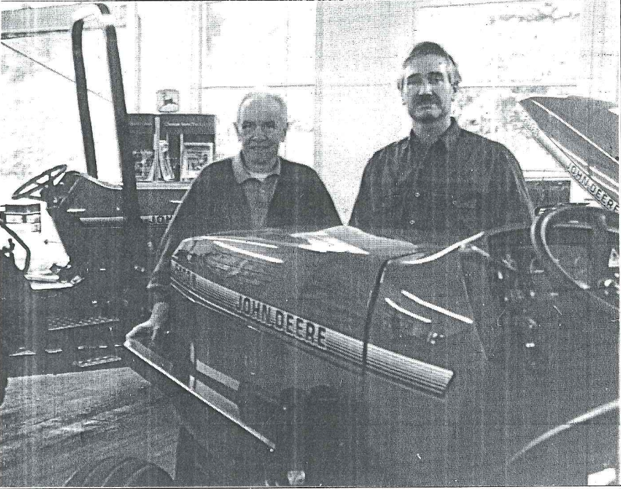 Two men standing behind a vintage race car inside a garage or workshop.