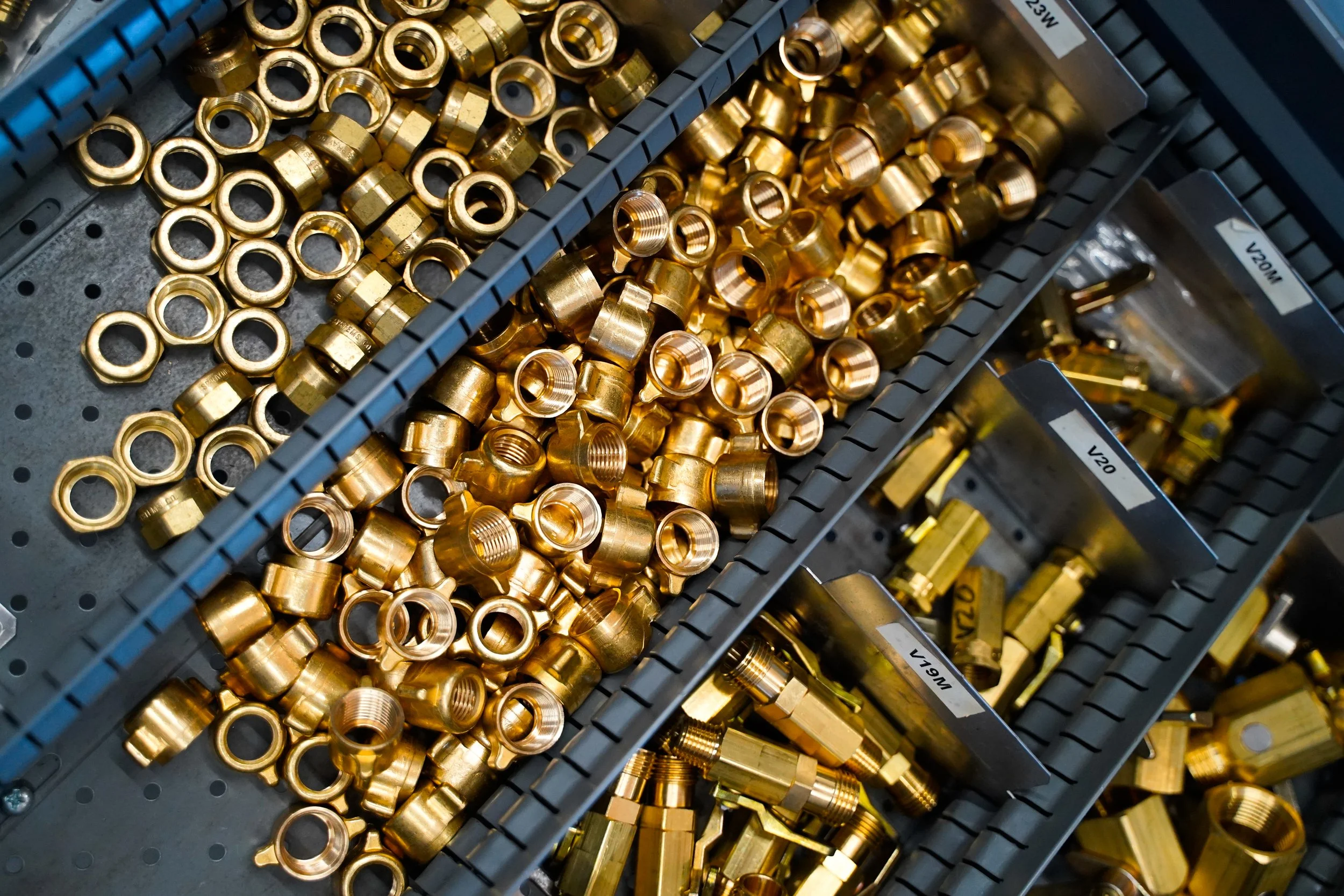 A bin filled with shiny, golden metal nuts and threaded fittings stored in partitions, likely used for industrial or mechanical purposes.