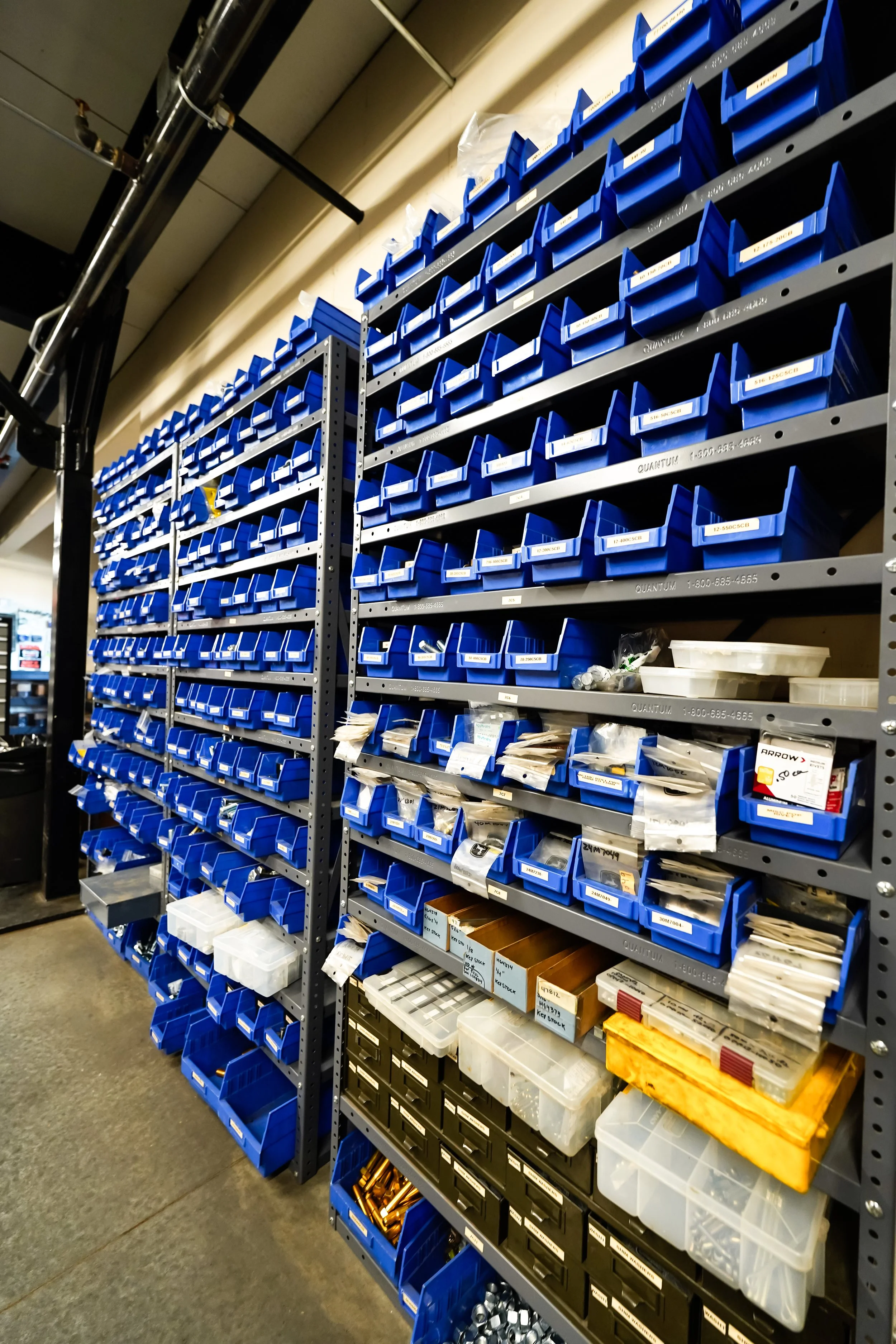 Industrial storage shelves filled with small plastic bins in blue, black, and clear, containing various nuts, bolts, and small hardware items in a workshop or warehouse.