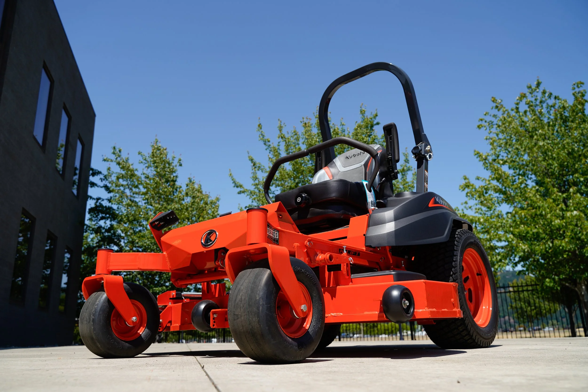 An orange riding lawn mower parked on a concrete surface with trees and a building in the background, taken from a low angle during the day.