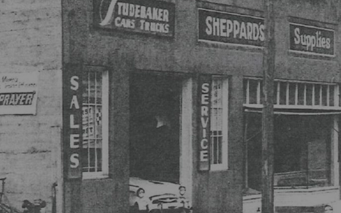 Black and white photo of storefront with signs reading "Tudekaser Cars Trucks," "Sheppard's," and "Supplies," with a small car parked in the entrance and vertical signs reading "SALES" and "SERVICE."