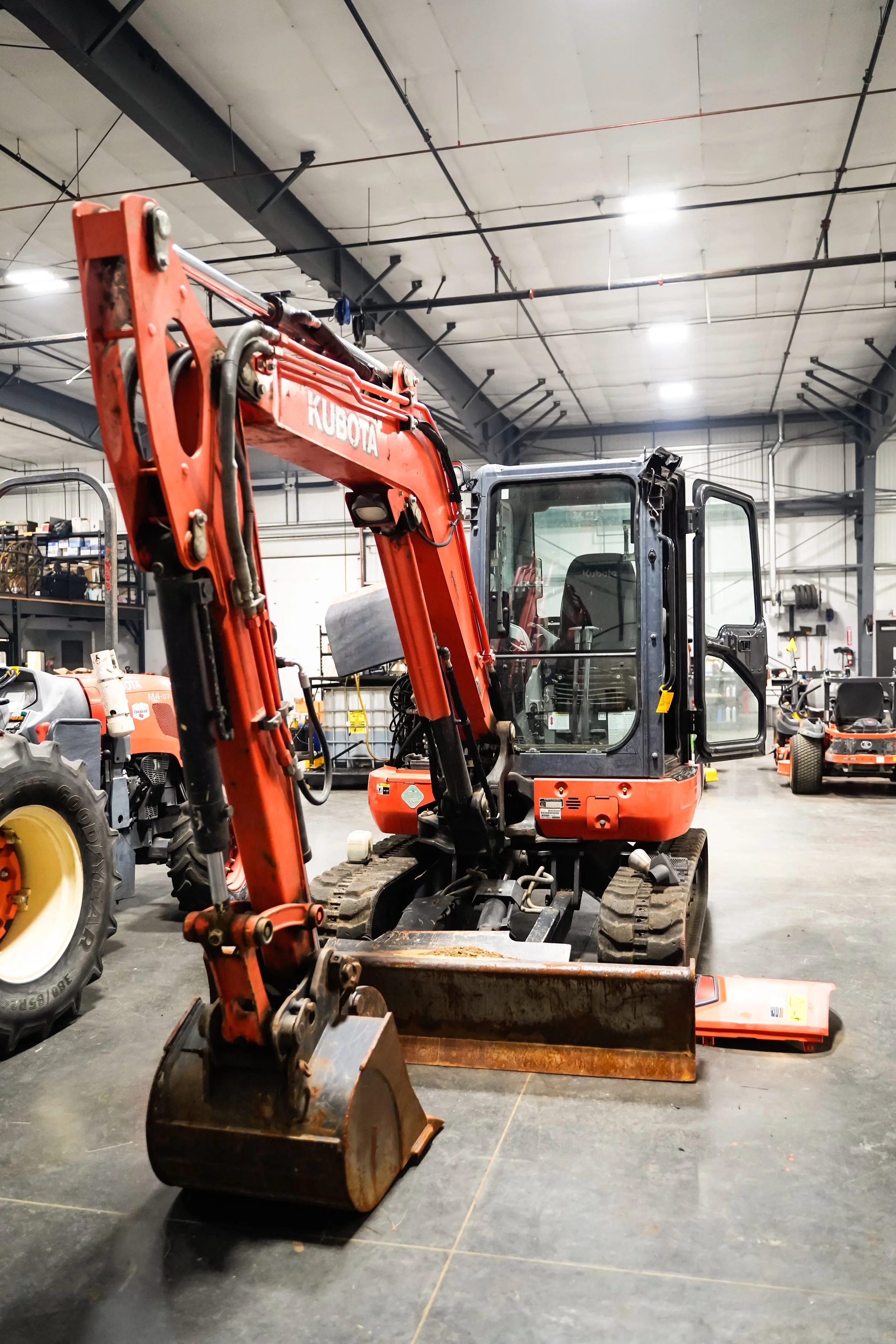 Indoor warehouse with construction equipment, including an orange Kubota mini excavator and other machinery, in a well-lit industrial space.