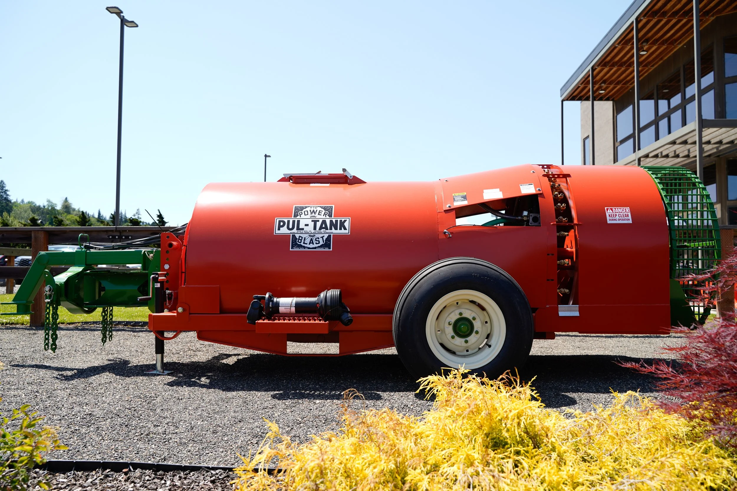 A large red and green power washer labeled 'Pul-Tank' is parked outdoors near buildings with large windows. There are trees and shrubs visible in the background.
