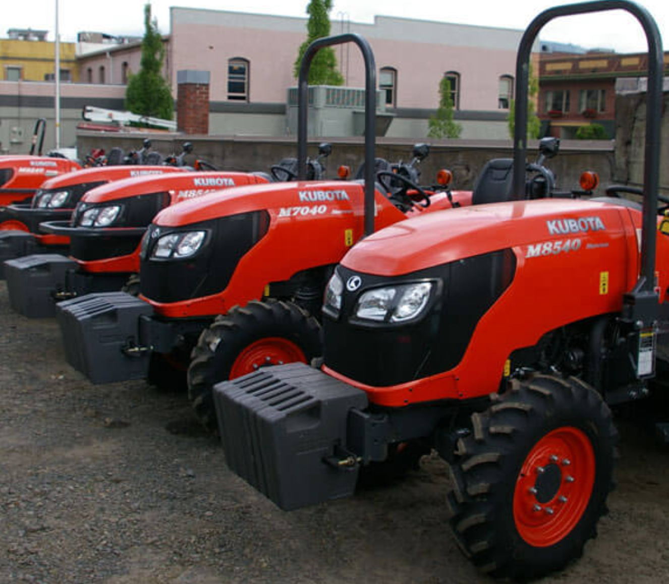 Multiple red Kubota tractors lined up outdoors, with buildings and trees in the background.