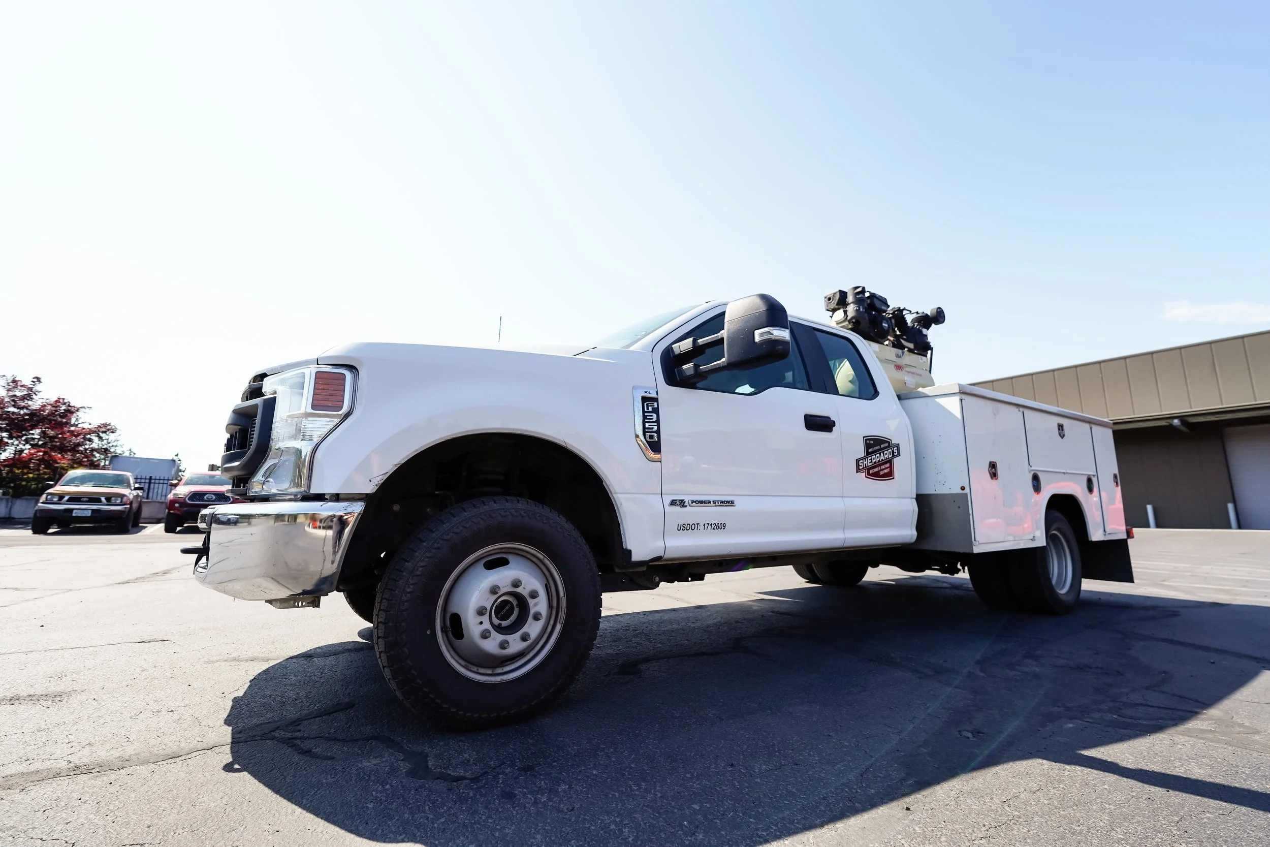 White utility truck with equipment on top parked in a lot with other vehicles and a building in the background against a clear blue sky.
