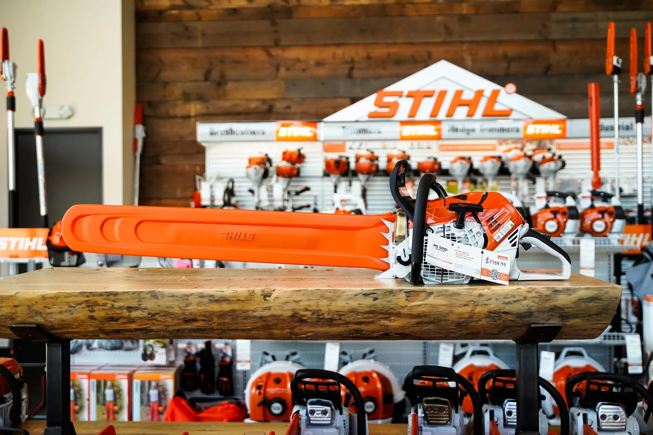 Chainsaw displayed on a wooden table inside a store, with shelves of power tools, helmets, and other equipment in the background.