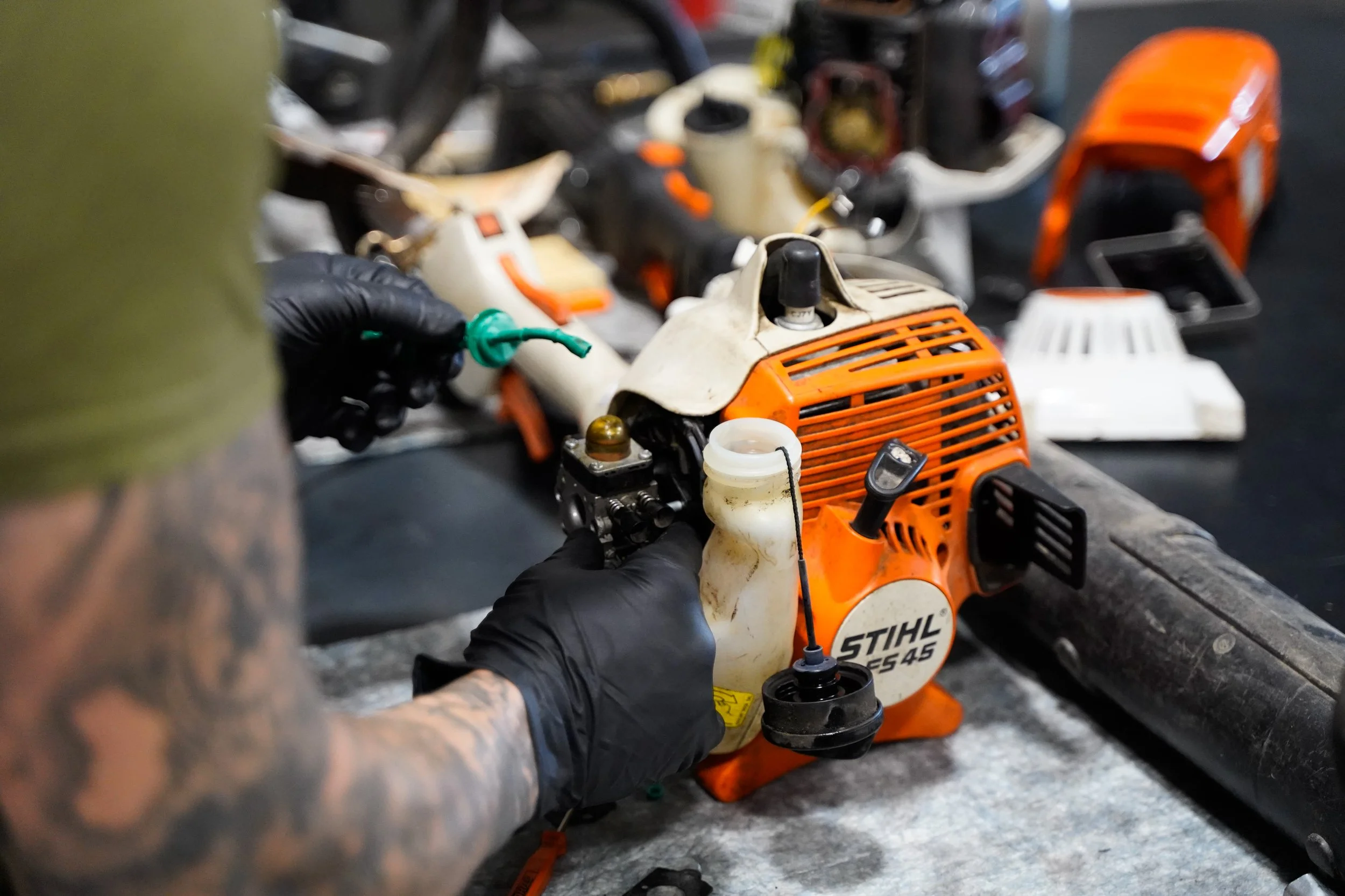 Person wearing black gloves repairing a small orange and white STIHL chainsaw, with various saw parts and tools on a workbench.
