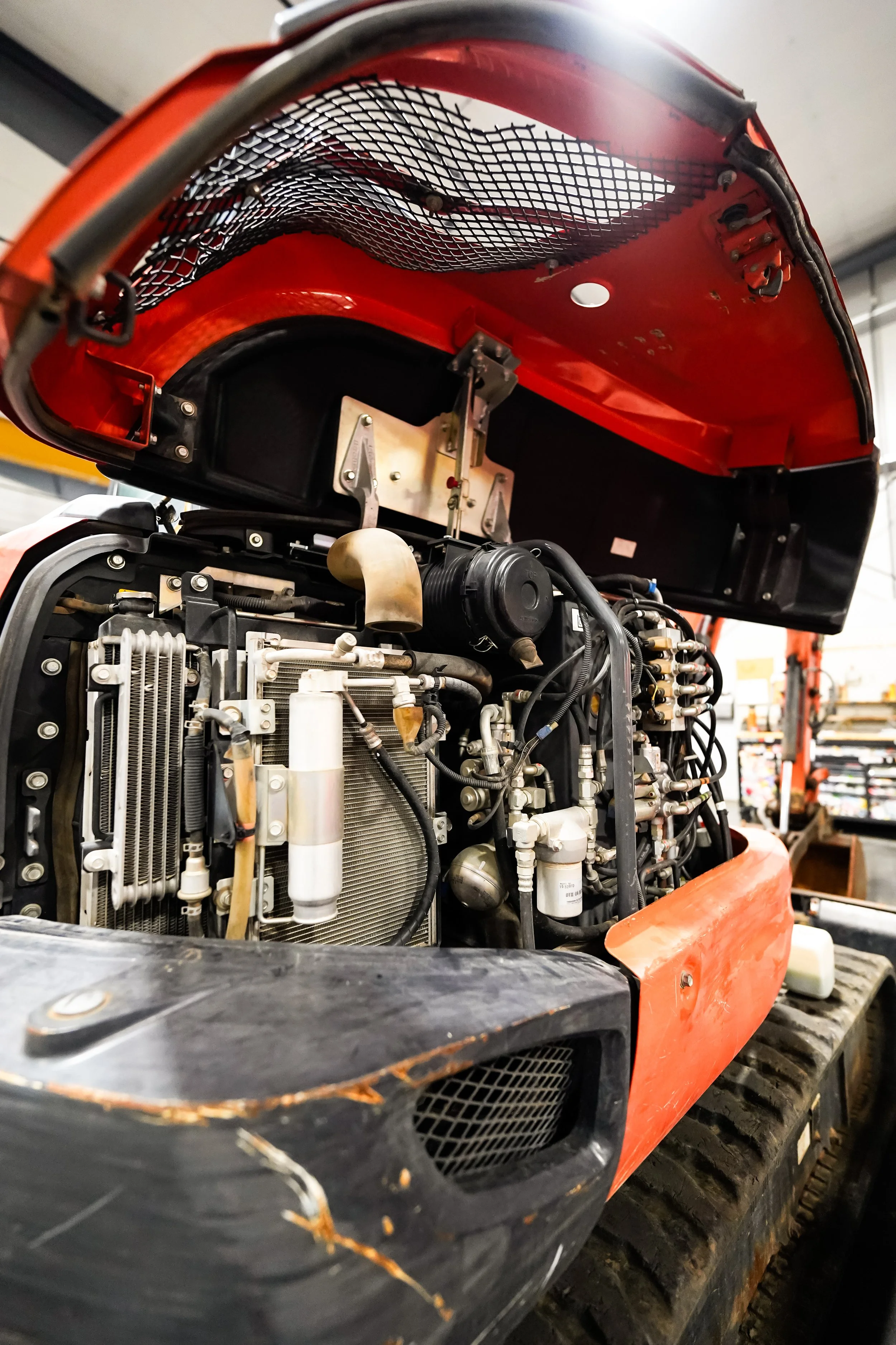 Close-up view of the engine of a construction or industrial machine, with exposed mechanical parts and hoses, under an orange cover with some damage.