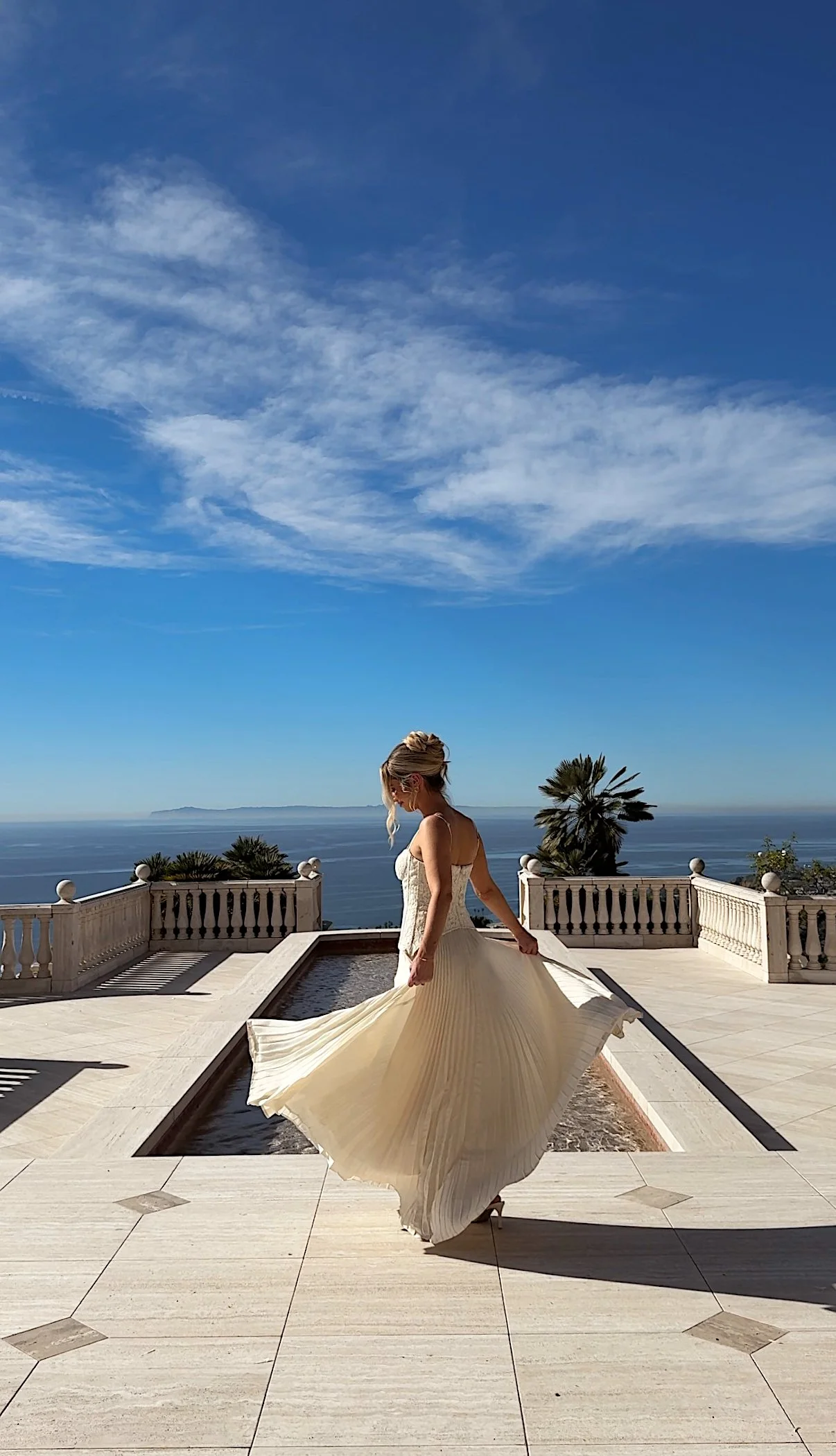 A woman in a beige wedding dress twirling on a balcony with a view of the ocean, palm trees, and a partly cloudy blue sky in the background.