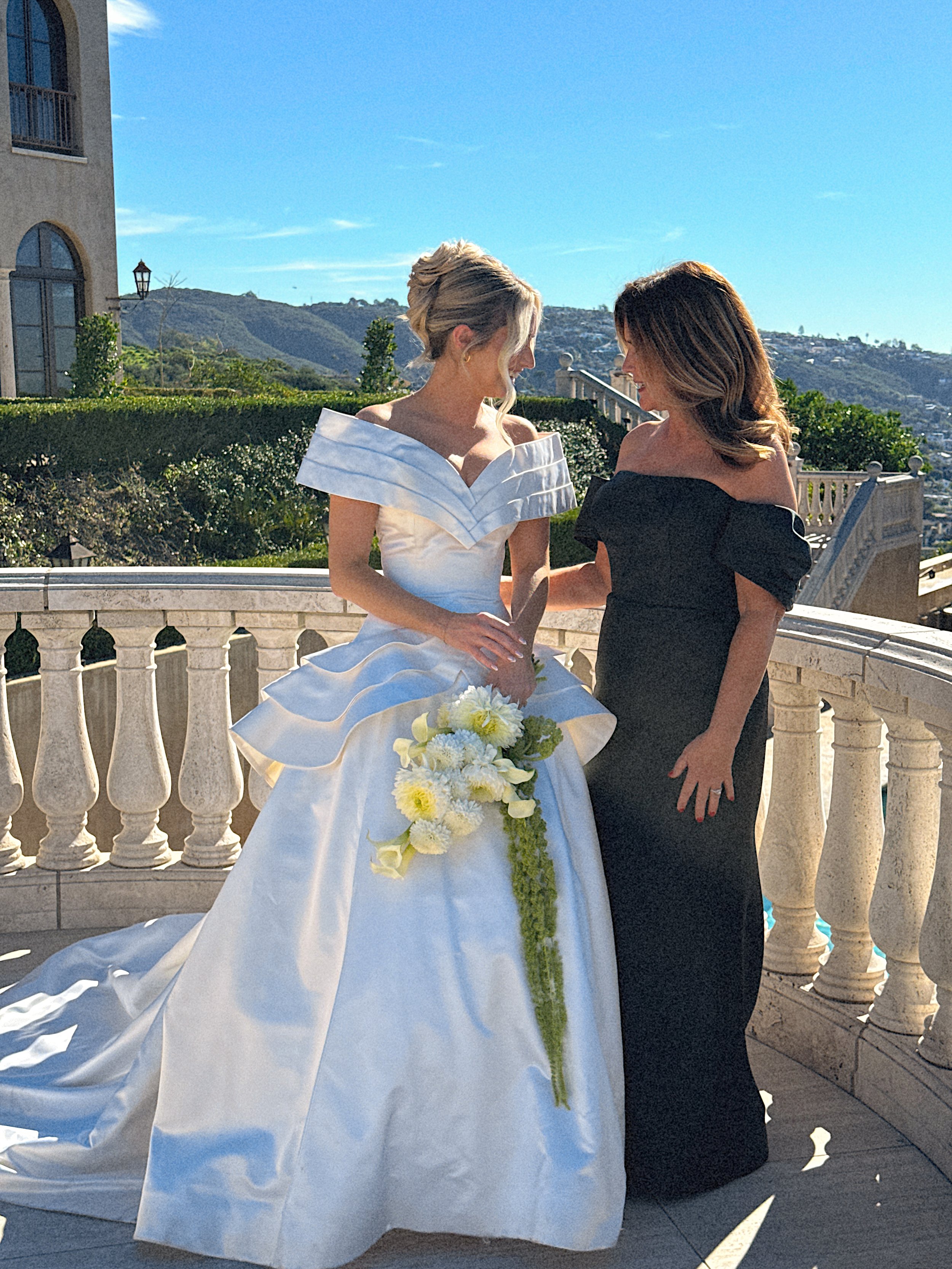 Two women standing on a balcony with a scenic landscape behind them, one in a white wedding dress holding a bouquet of white and yellow flowers, the other in a black dress, smiling at each other.