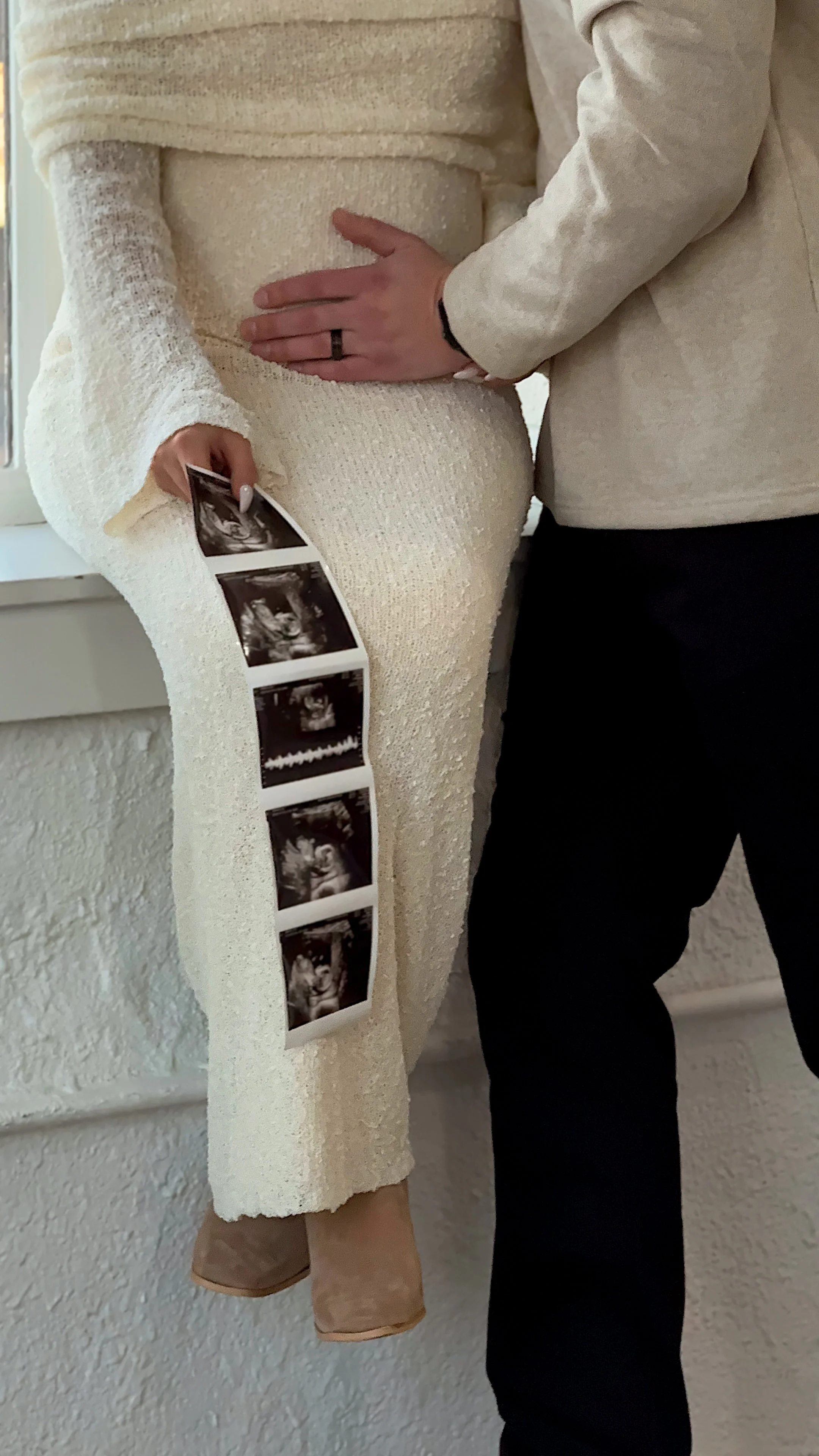 Pregnant woman in a cream-colored dress holding ultrasound images, with her partner's hand on her belly. The woman is wearing a long-sleeved dress, and the partner is dressed in a light-colored shirt and dark pants. They are standing against a white textured wall.