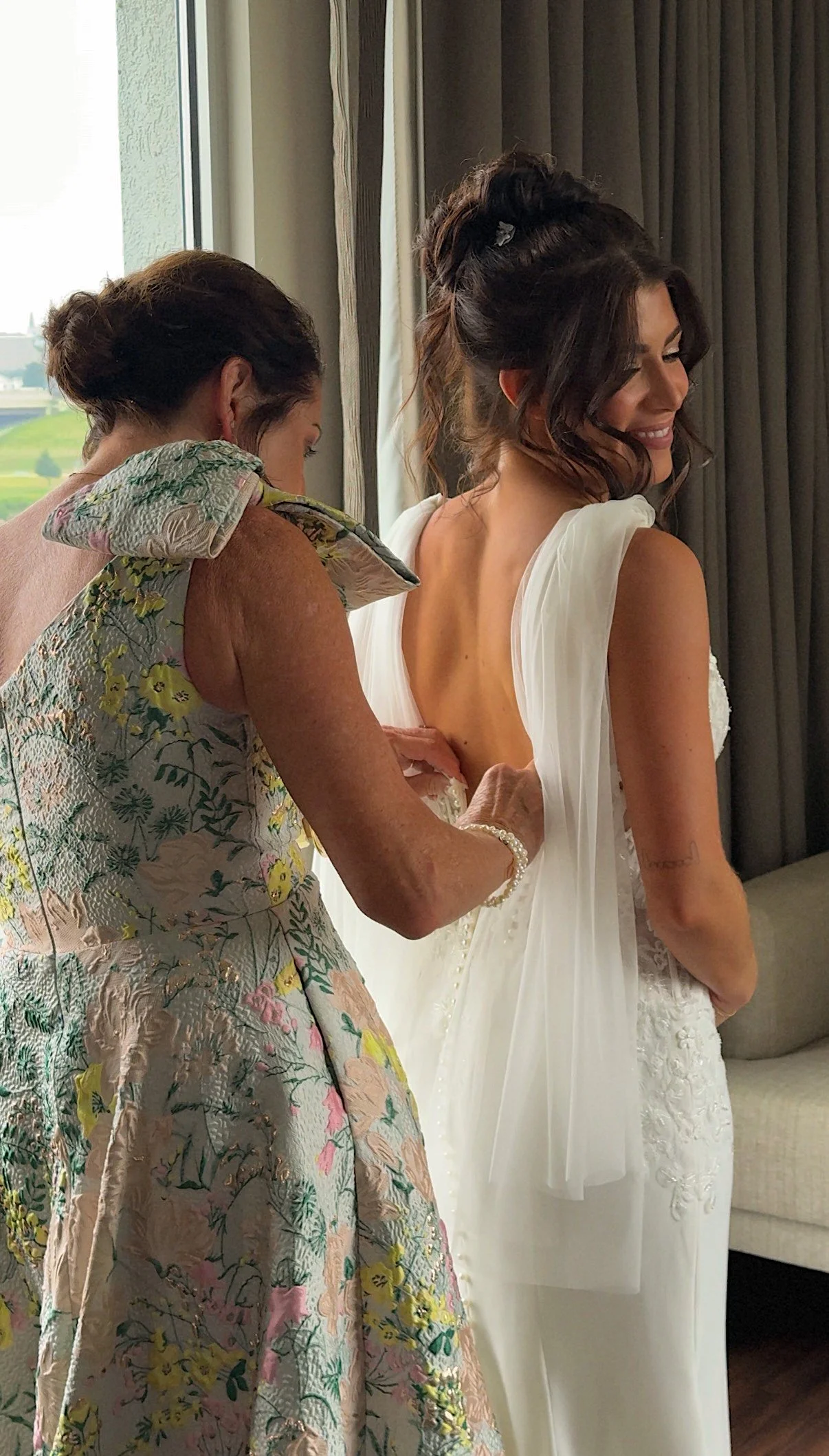 A woman in a white wedding dress is being assisted by another woman in a floral dress, likely helping her get ready for her wedding.