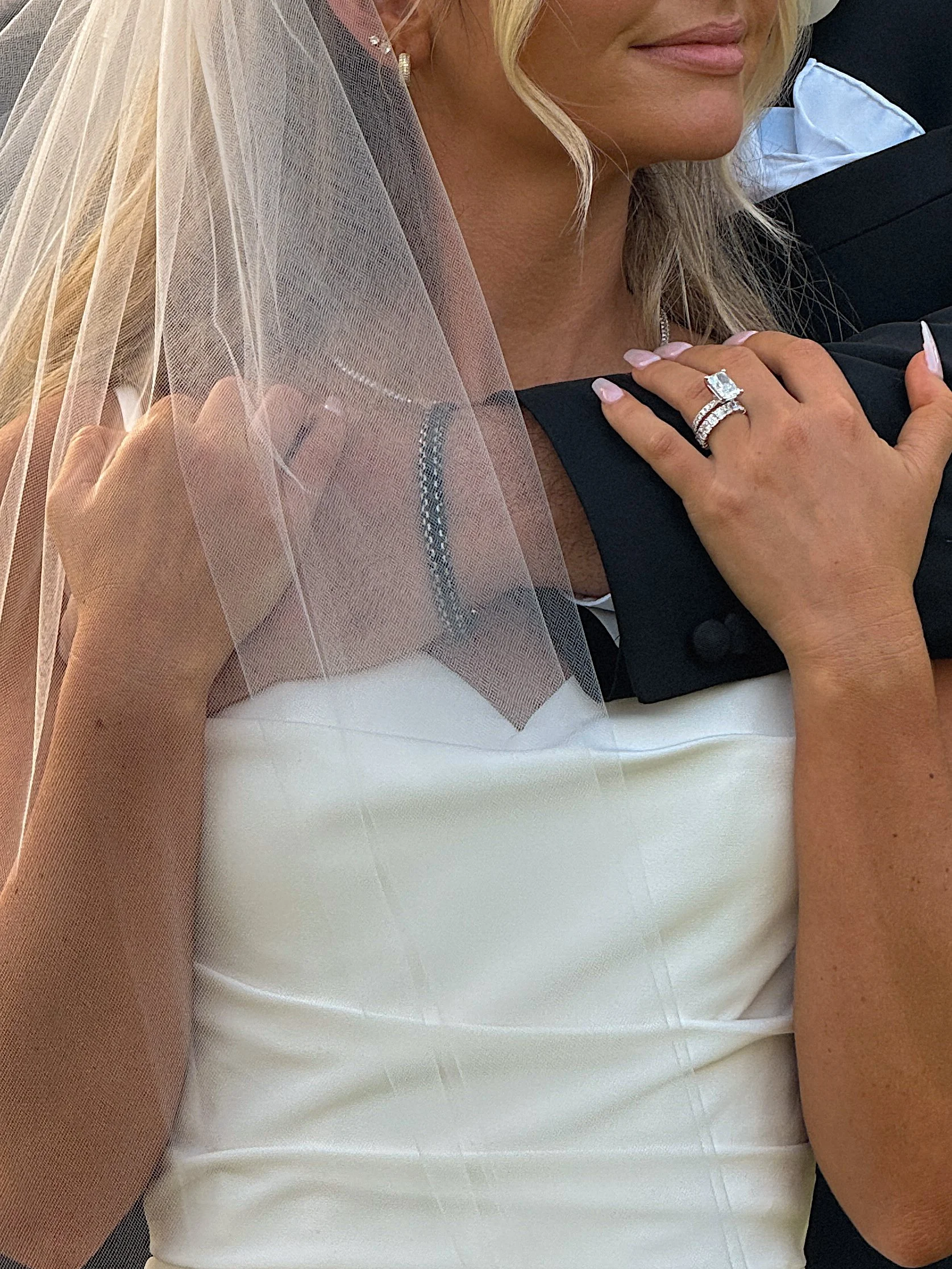 A woman with blonde hair, wearing a white dress, a wedding veil, and jewelry, sitting close to a man in a black suit, during a wedding or wedding reception.