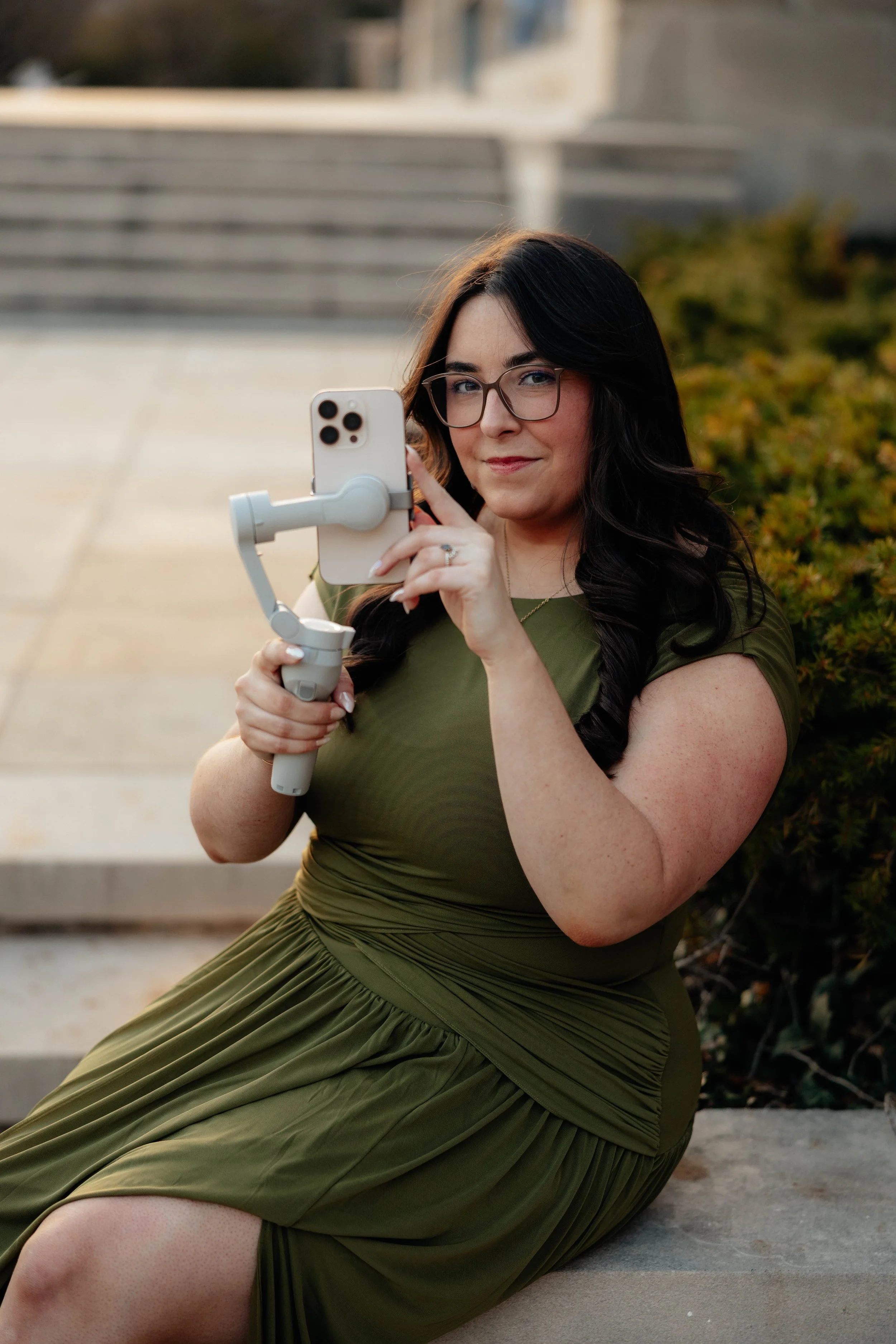 A woman with long dark hair, glasses, wearing a green dress, is sitting on a concrete surface outdoors, holding and looking at her smartphone mounted on a stabilizer.