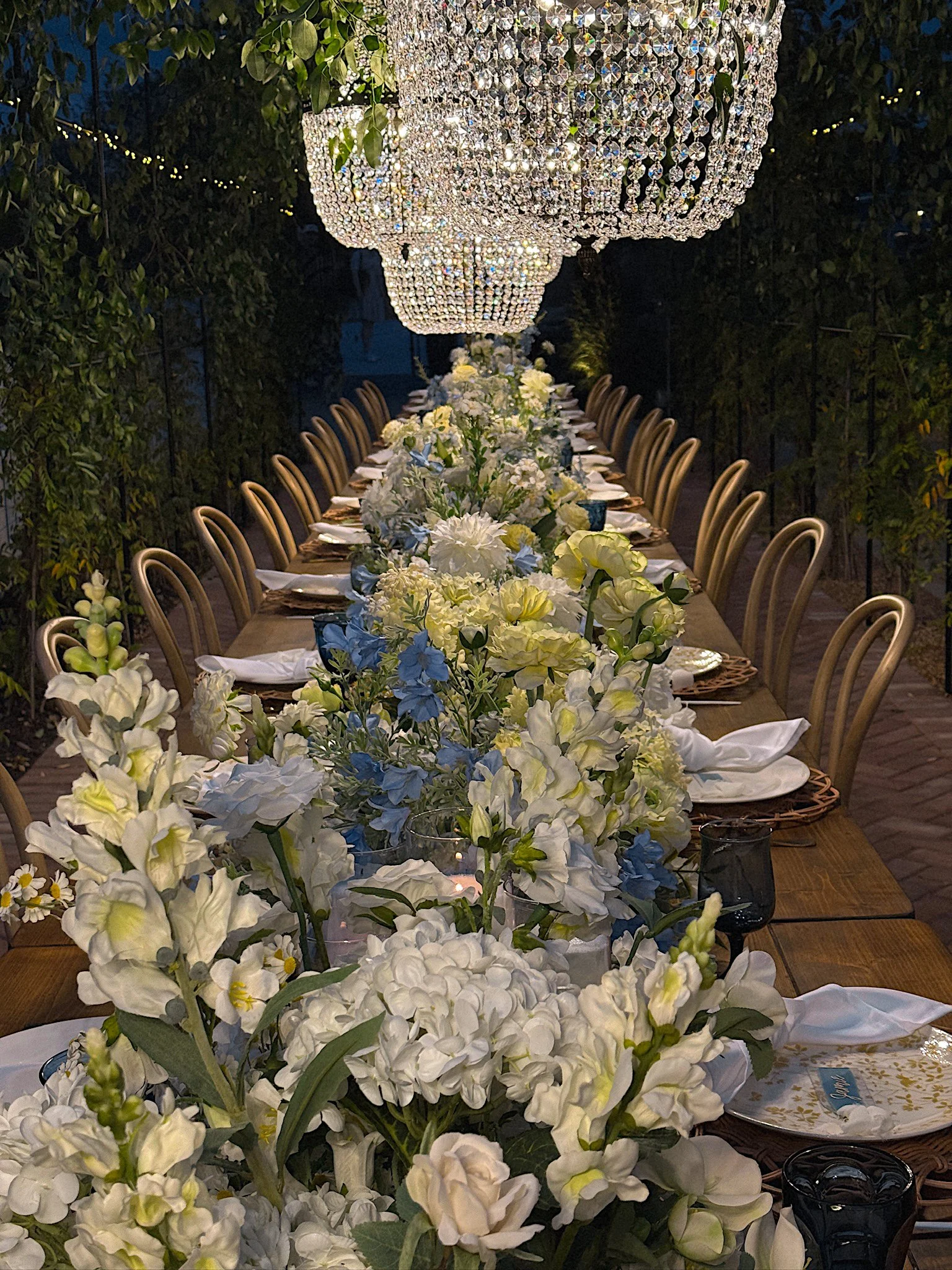Elegant outdoor event table decorated with white, yellow, and blue flowers, set with plates, napkins, and glasses, under large crystal chandeliers at night.