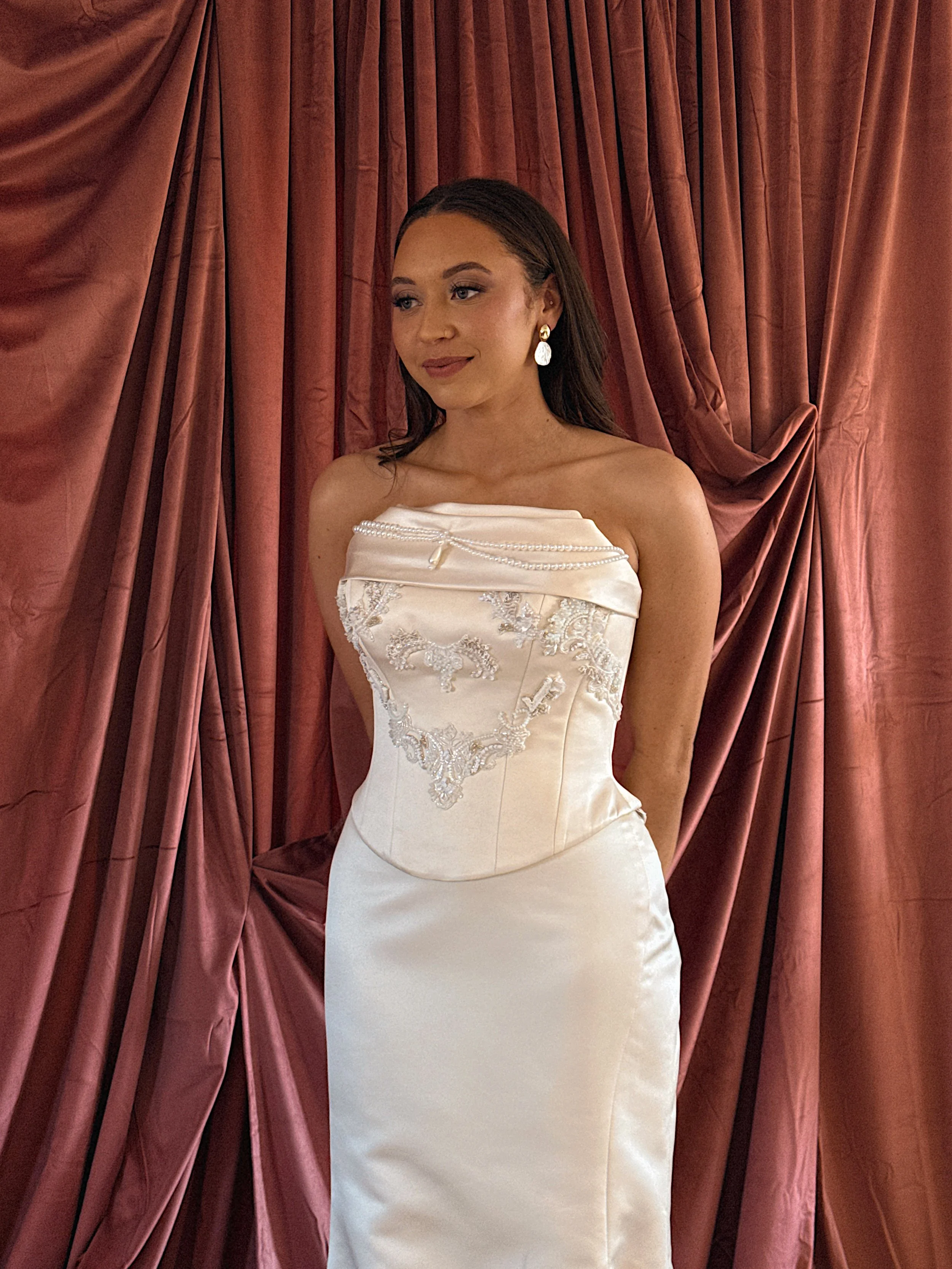 A woman in a white lace wedding gown with pearl and gold earrings standing in front of a red velvet curtain.