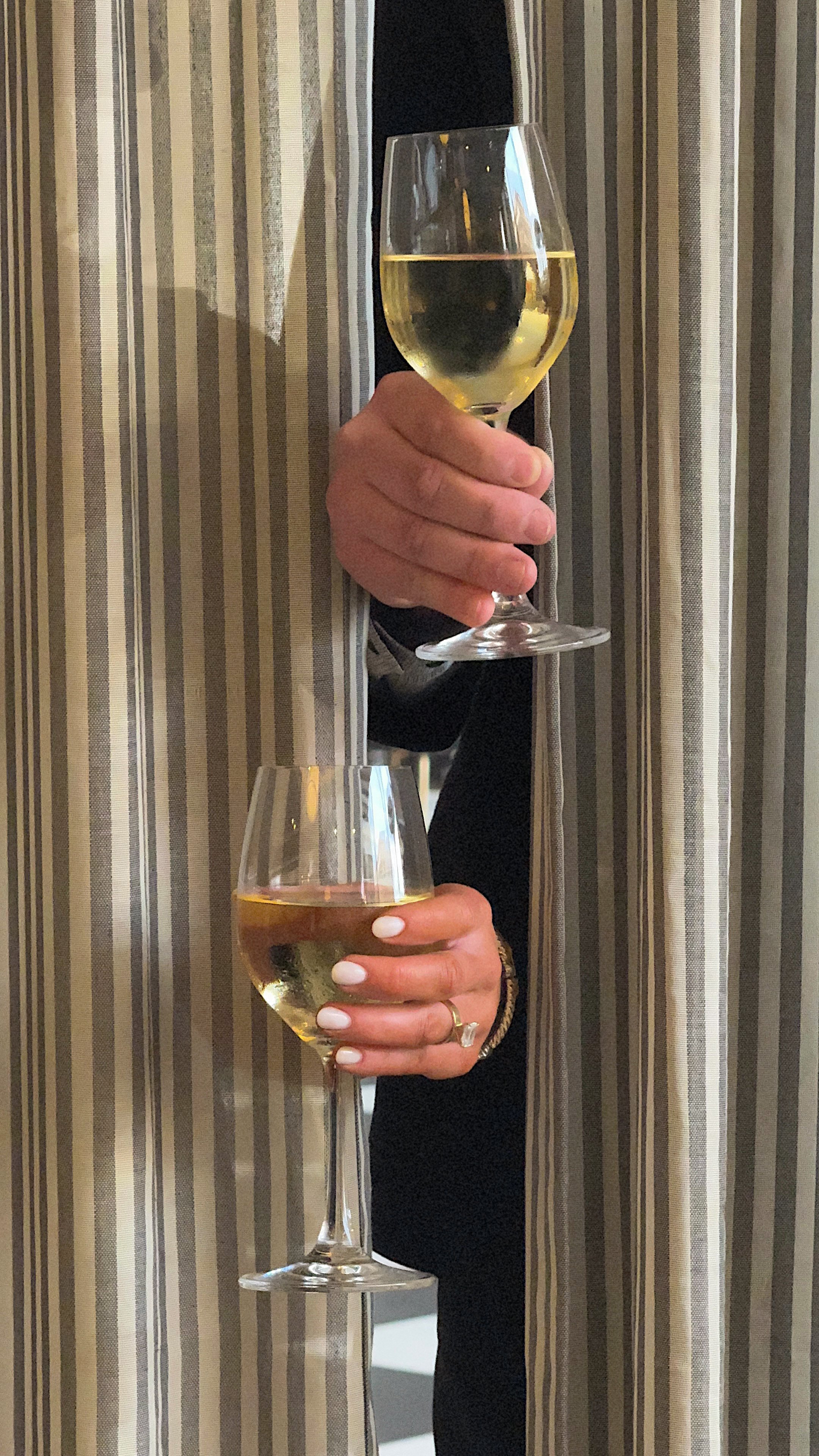Two people holding glasses of white wine at a social event, with one person's hand visible and the other partially visible behind a striped curtain.